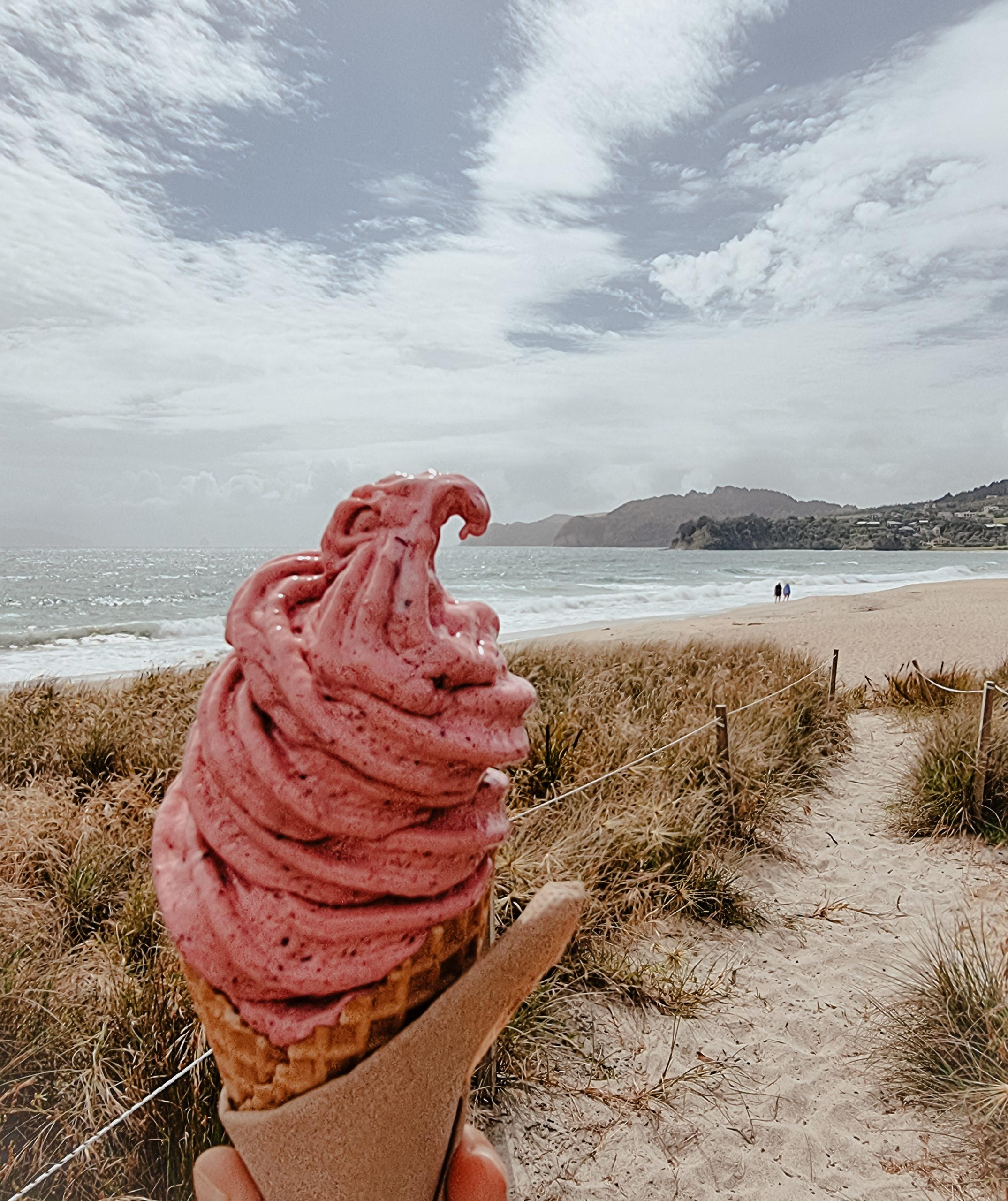 Hand holding a cone with pink ice cream in front of a beach - Ice cream