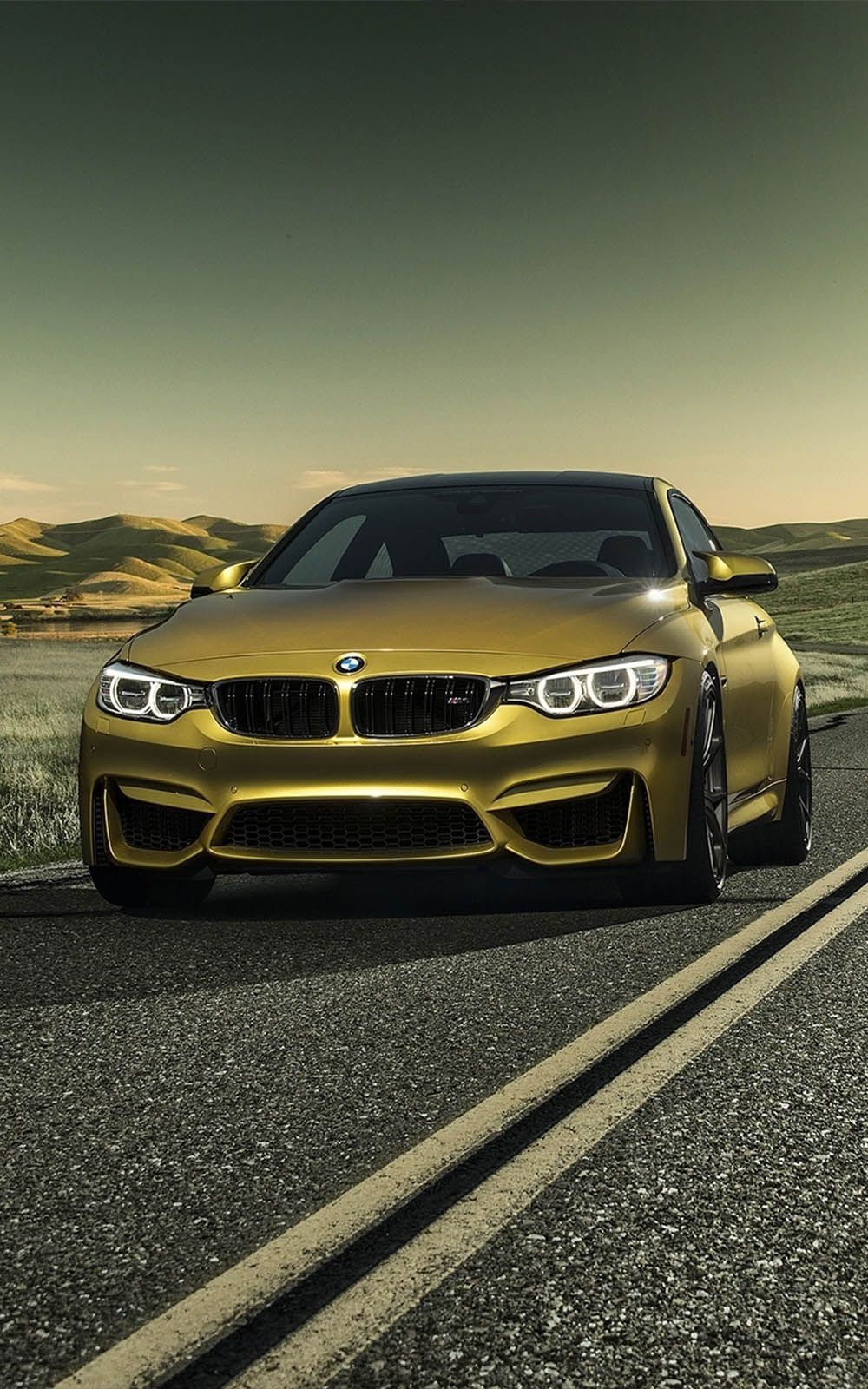 A gold BMW M4 sits on the road with the sun setting in the background. - BMW