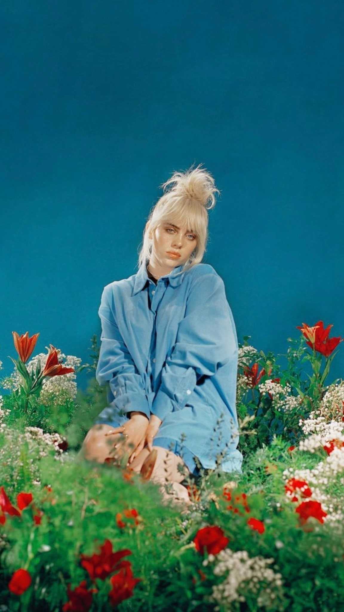 Billie Eilish in a blue shirt sitting in a field of flowers - Billie Eilish