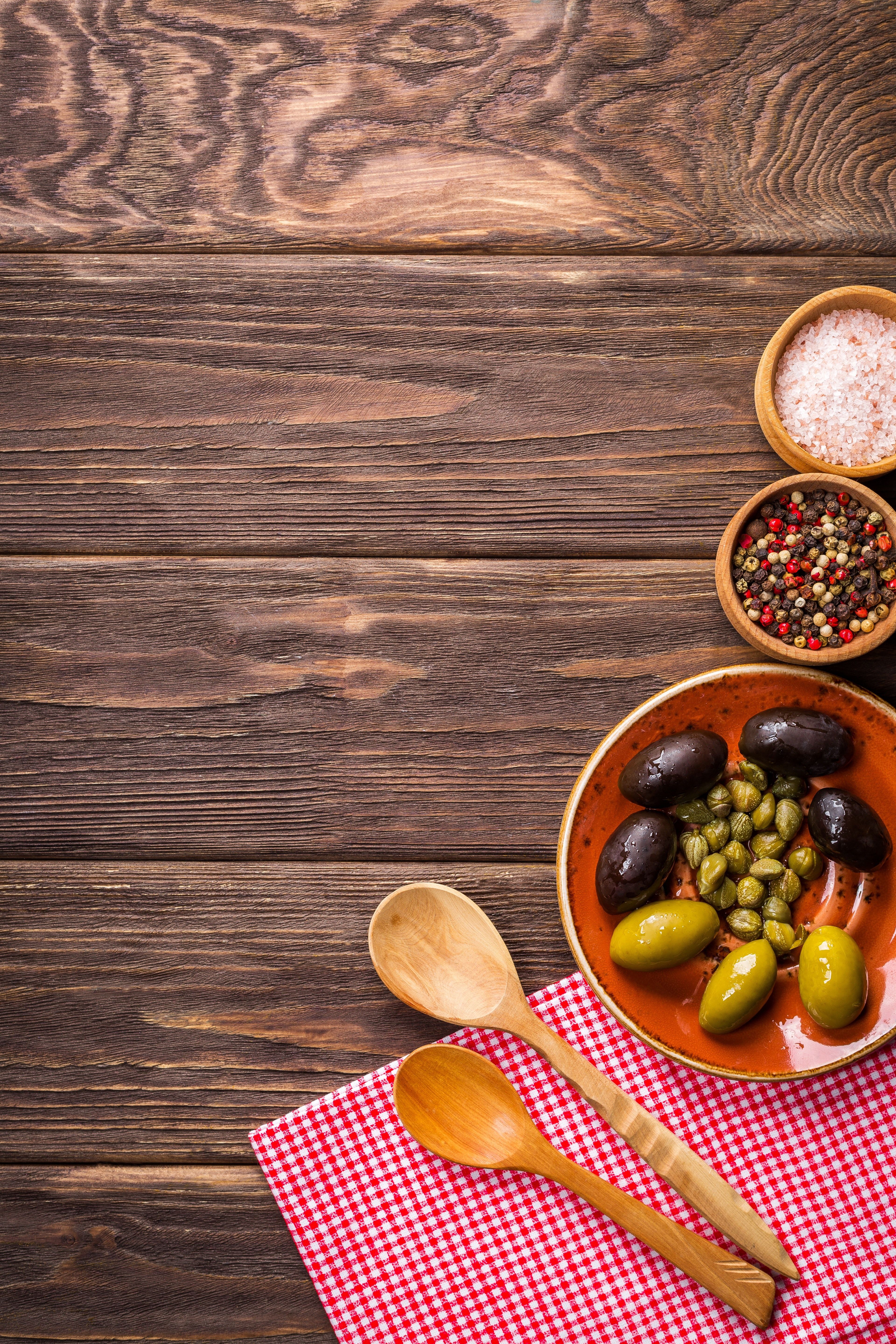 A wooden table with a plate of olives and salt, pepper and a napkin with spoons. - Food