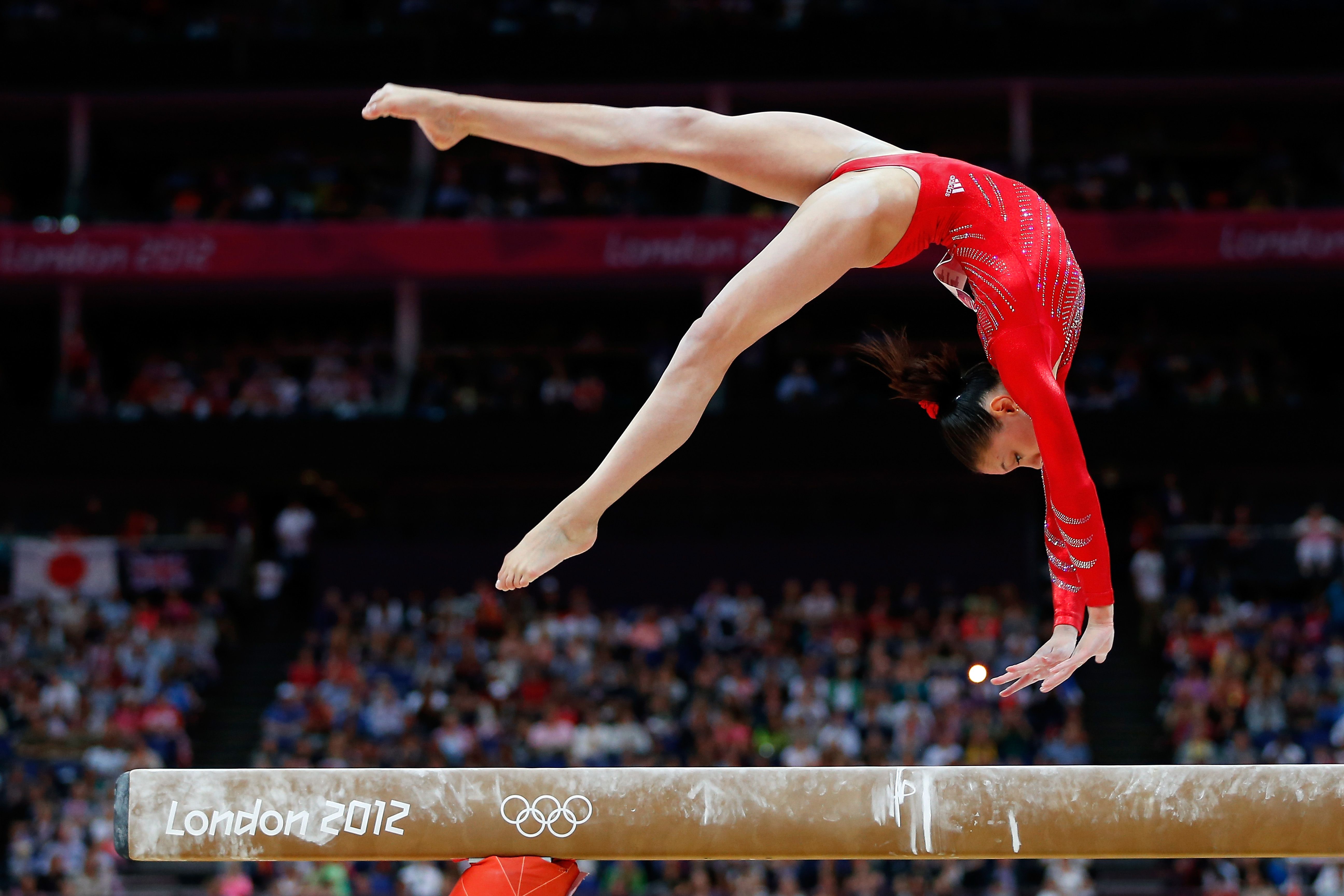 A gymnast in a red leotard is mid-air on a balance beam. - Gymnastics