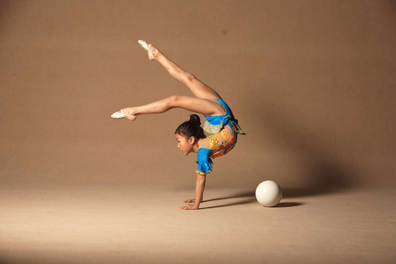 A young girl in a blue leotard doing a handstand next to a white ball. - Gymnastics