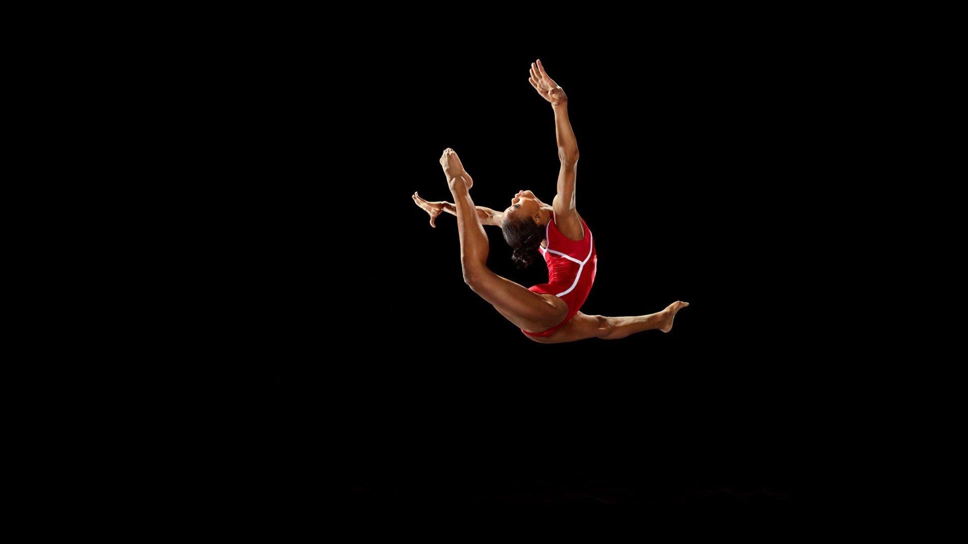 A woman in a red leotard and white shorts, with her arms raised, leaps through the air. - Gymnastics