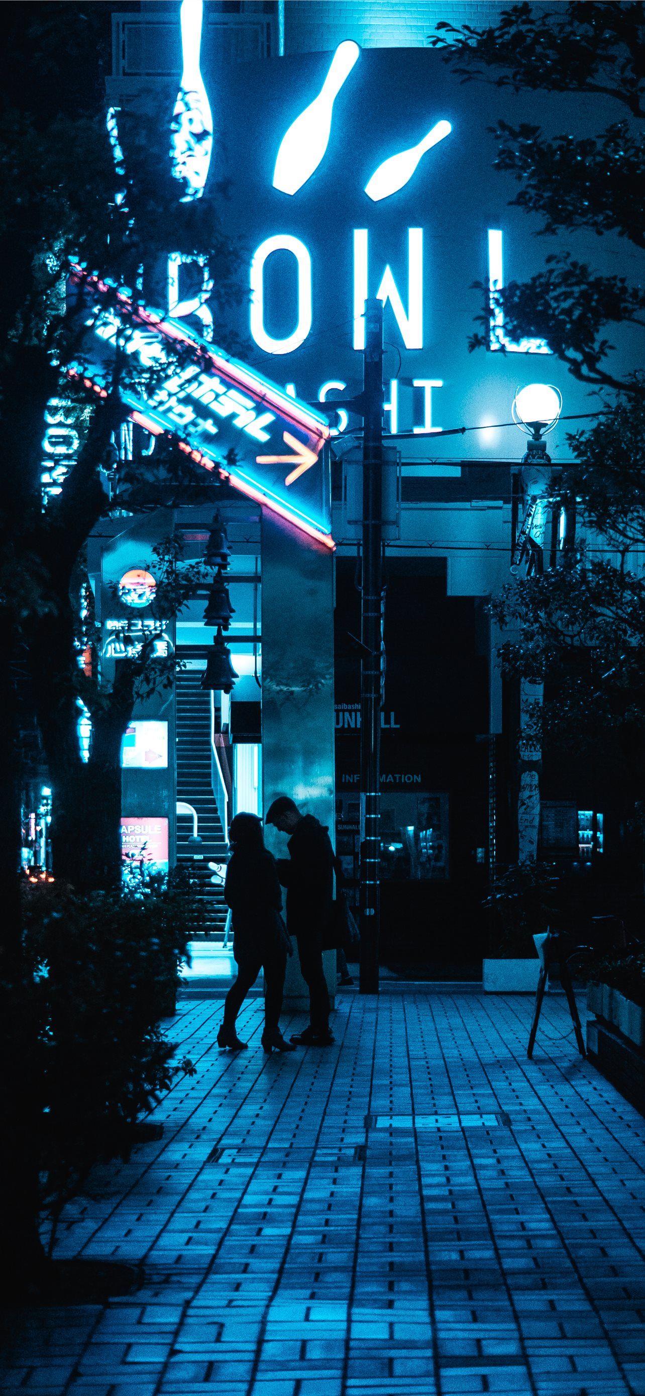 A couple walks down a street at night with a neon sign in the background. - Japan
