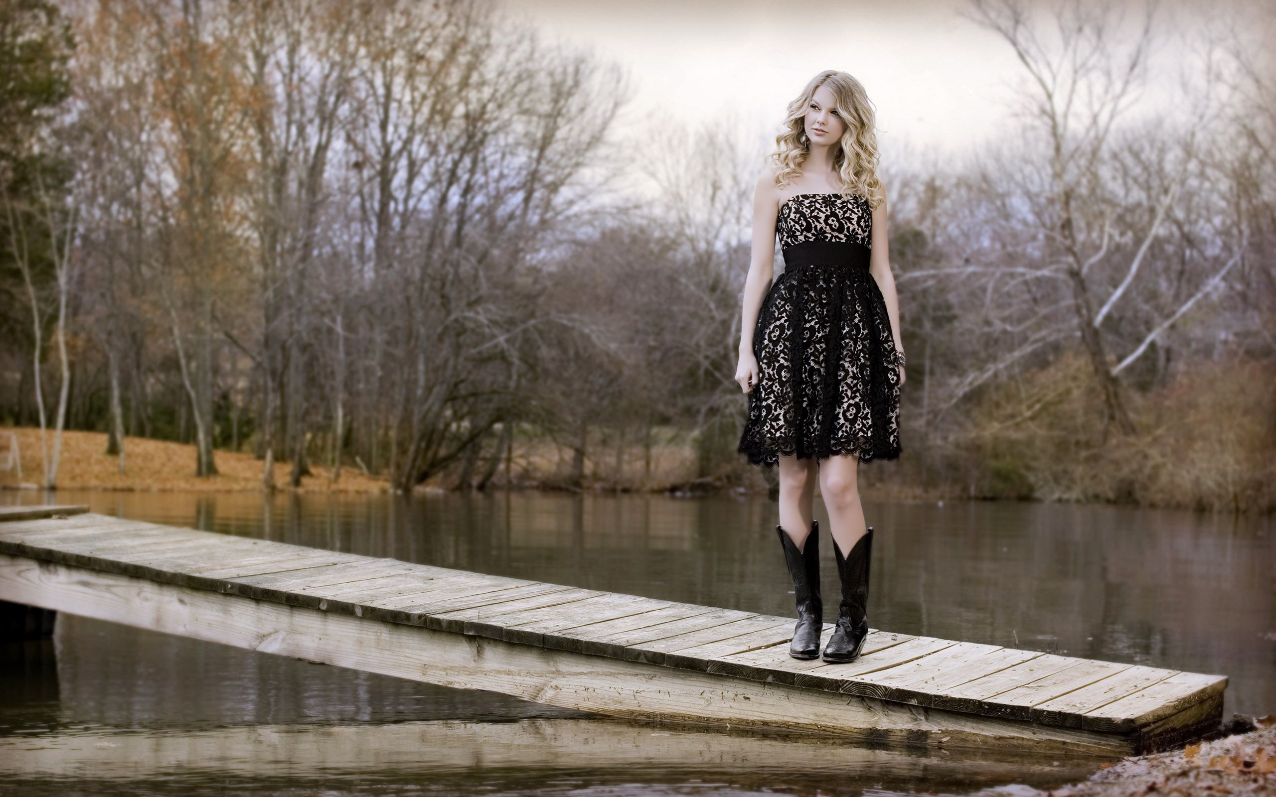 A woman in a black dress and cowboy boots stands on a dock. - Taylor Swift
