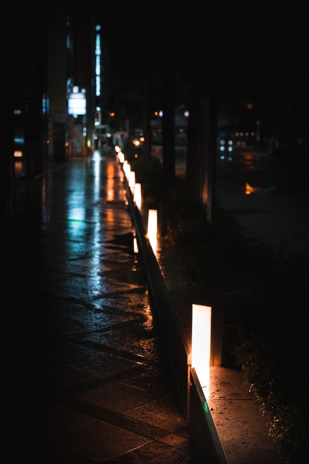 A row of lit posts along a wet sidewalk at night. - Night