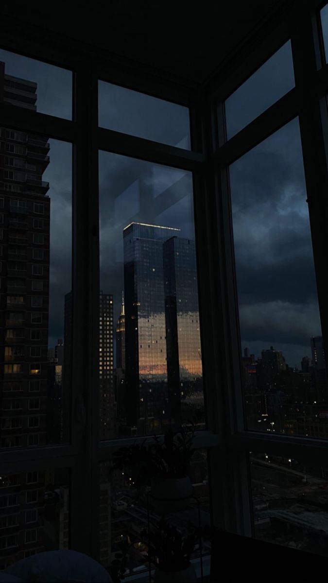 The sun sets behind a skyscraper as seen from a window in a city apartment. - Night