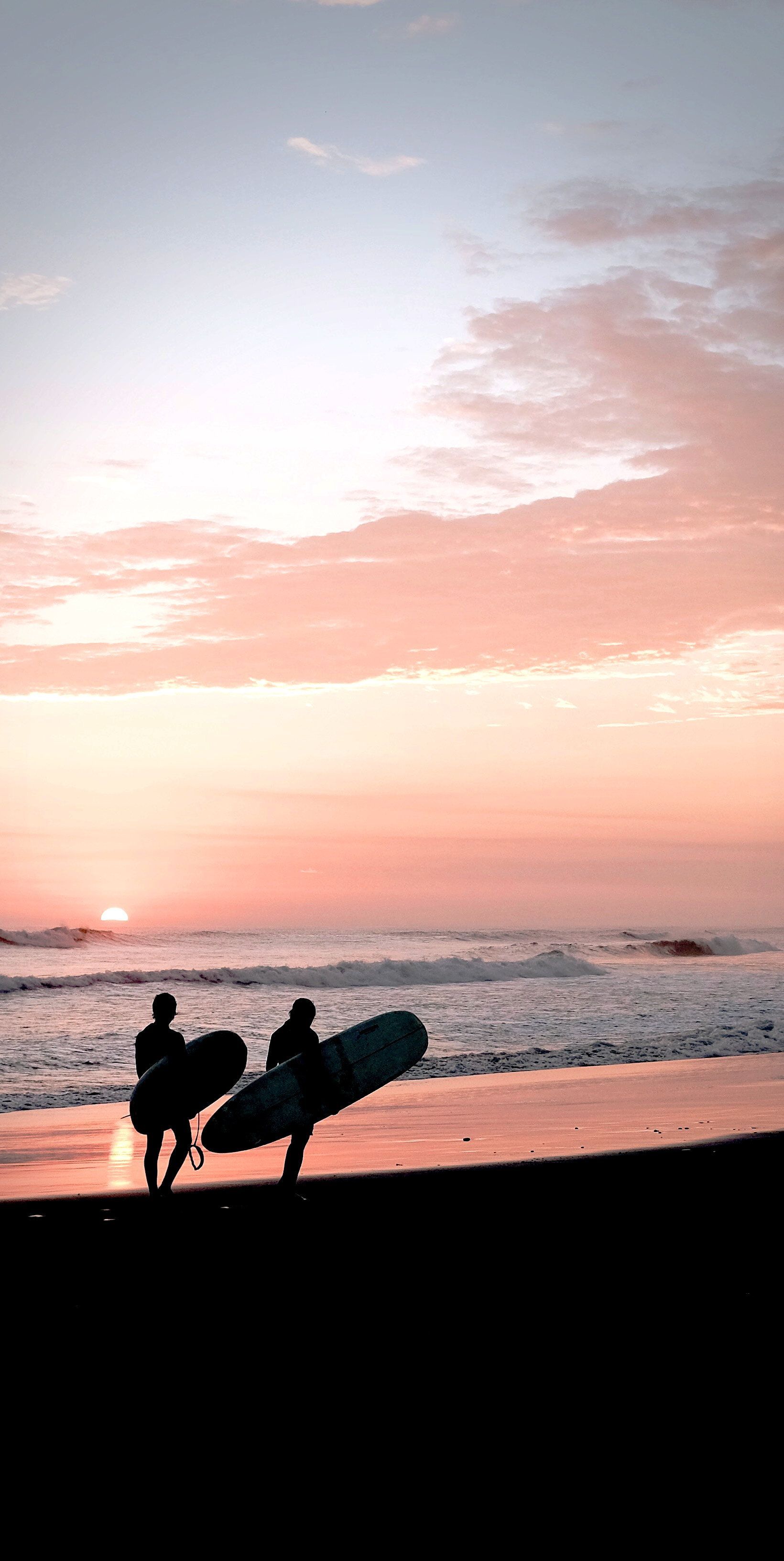 Two surfers carrying their boards along the beach at sunset. - Surf