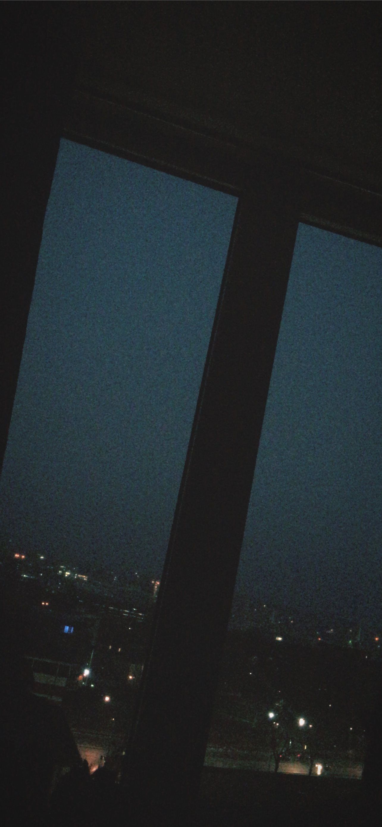 A dark room with two windows, one on the left and one on the right. The left window is covered with a blind. The right window shows a city skyline at night. - Lo fi