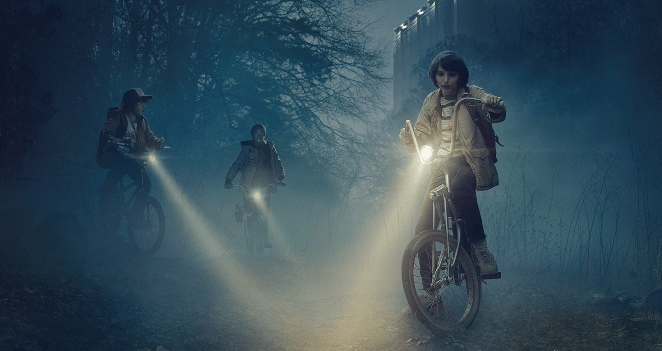 Three boys on bikes in a dark forest with one boy giving a thumbs up. - Netflix