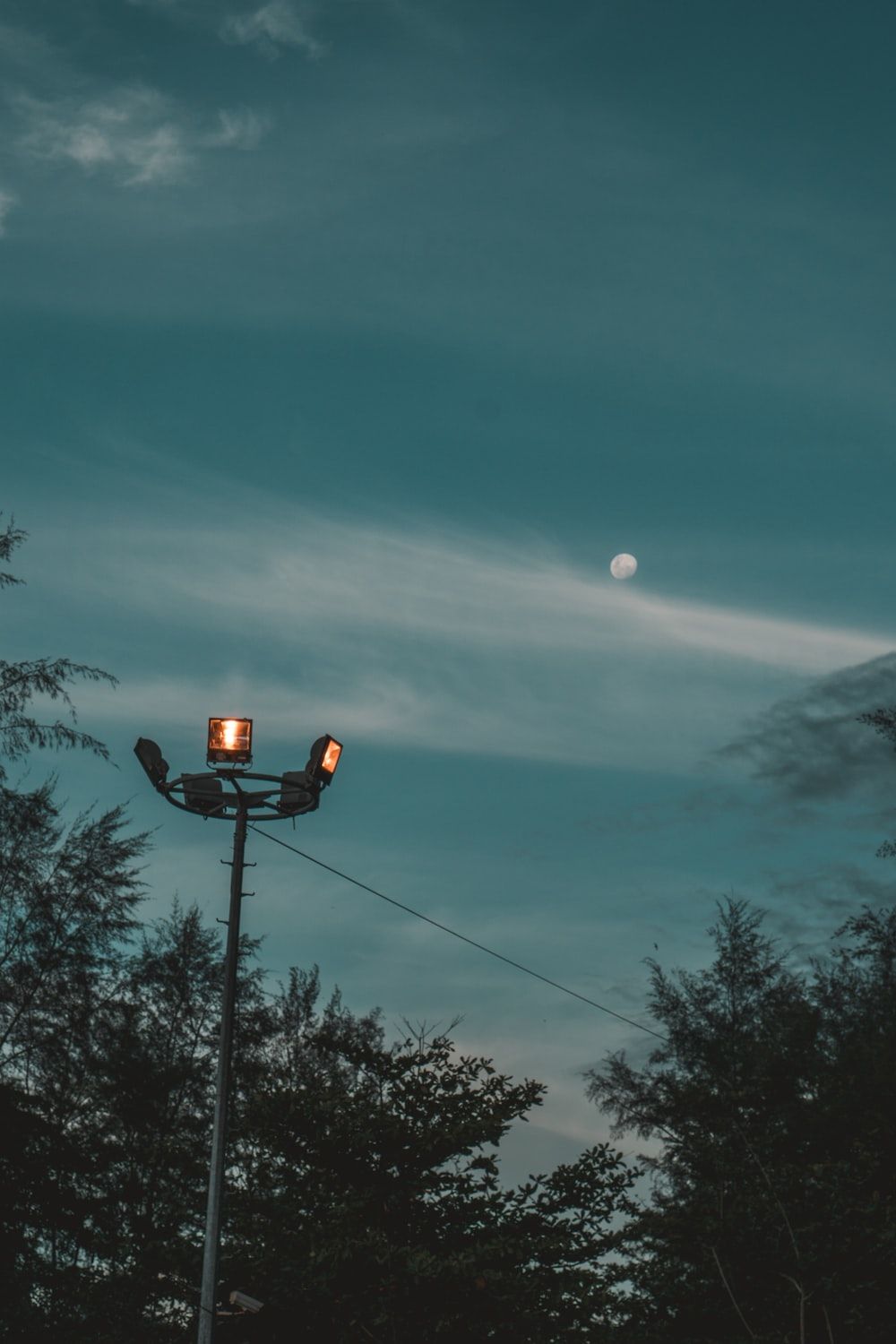 A streetlight with three lights on it, with a dark blue sky and the moon in the background.  - Twilight