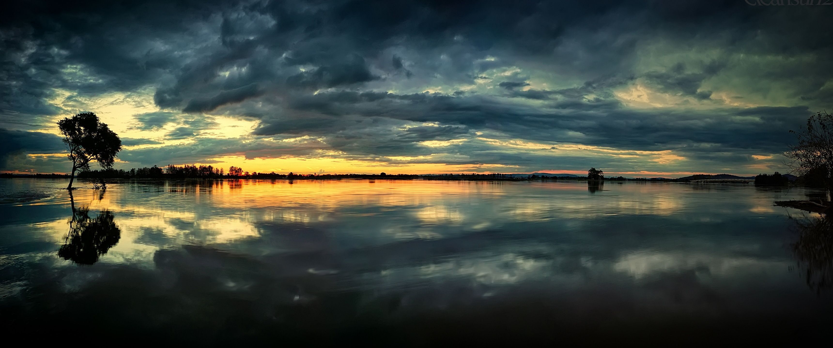 A dramatic sunset over a lake with a tree in the middle. - 3440x1440