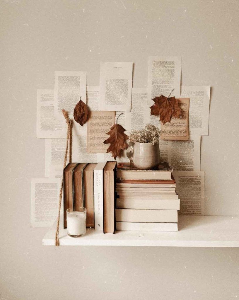 A bookshelf with a stack of books, a candle, and some dried leaves. - Light academia