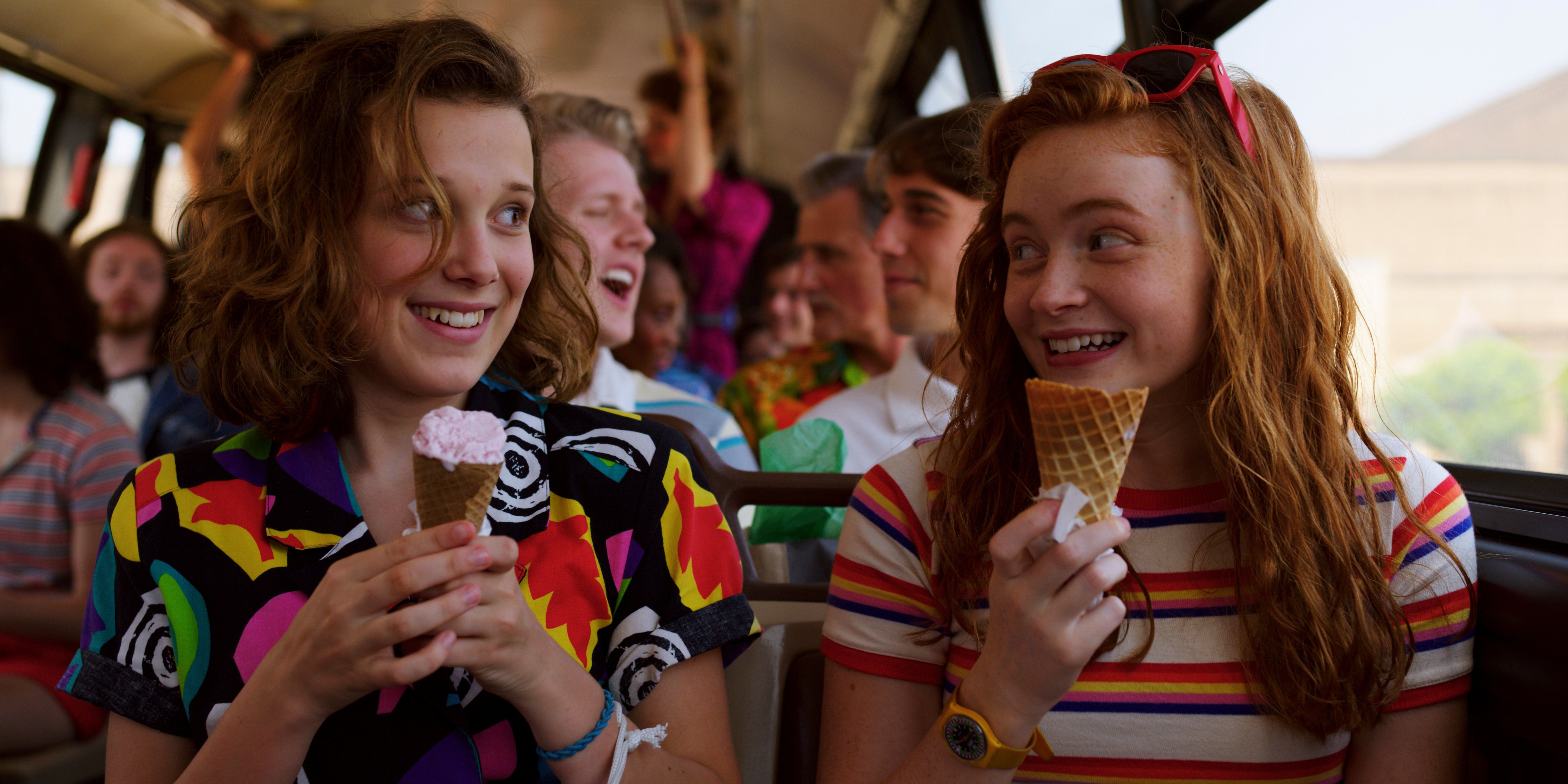 Sadie Sink and Millie Bobby Brown share a smile while eating ice cream on a bus in Stranger Things. - Stranger Things