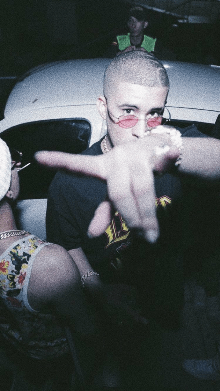 A man with a shaved head and pink glasses sitting in a car - Bad Bunny