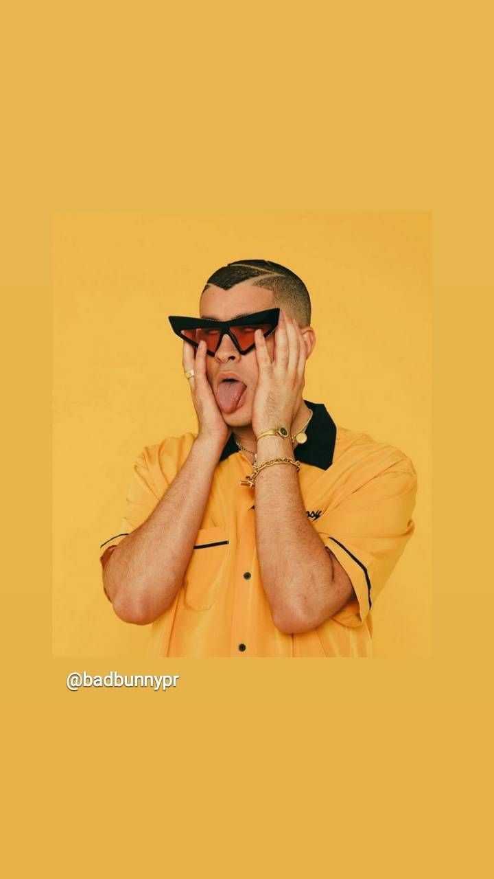 A man with a buzz cut and dark sunglasses covers his face with his hands against a yellow background. - Bad Bunny