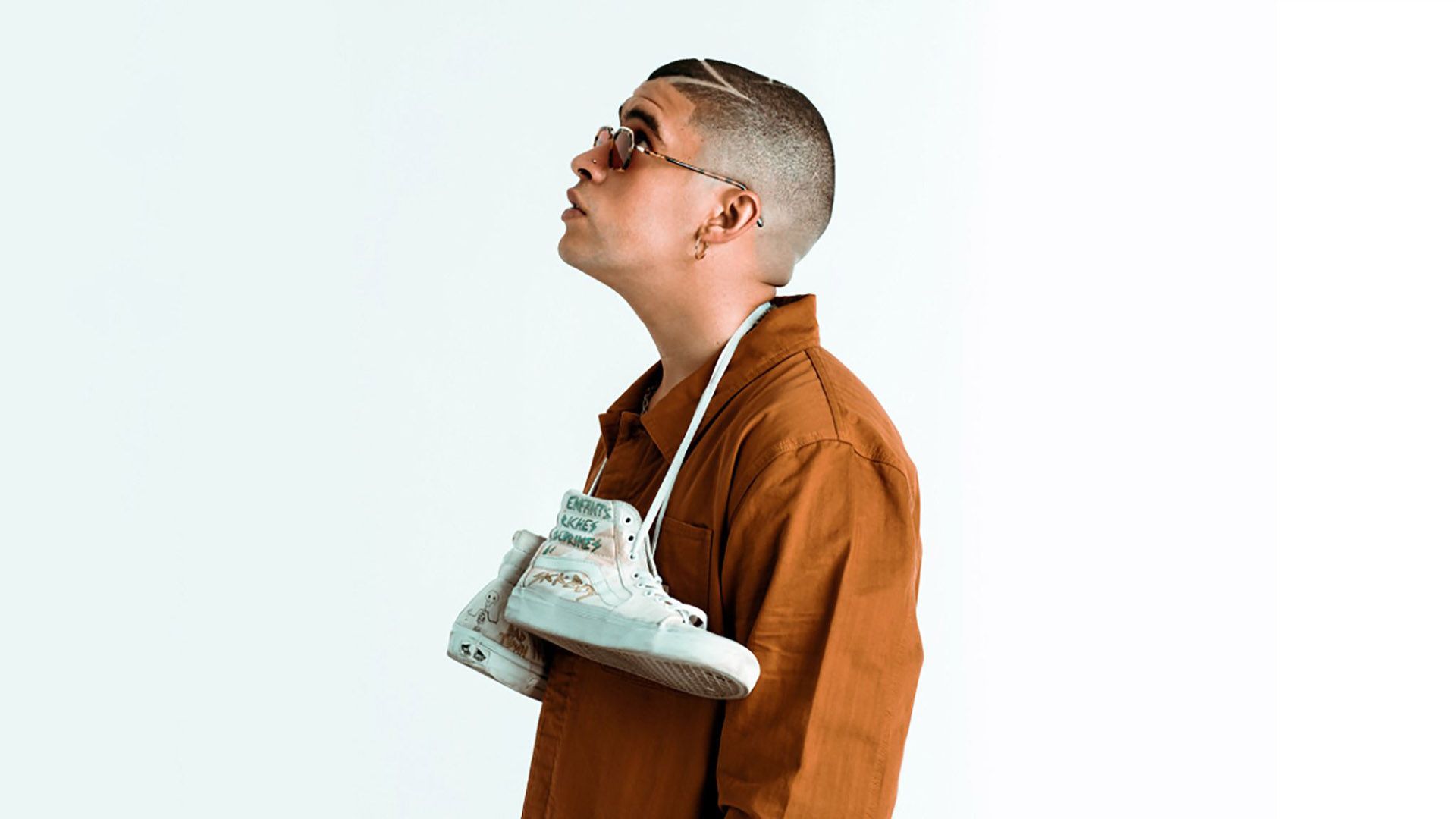 Bad Bunny Aesthetic With Hanging Shoes On Neck Looking Up Wearing Brown Coat In White Background HD Music Wallpaper