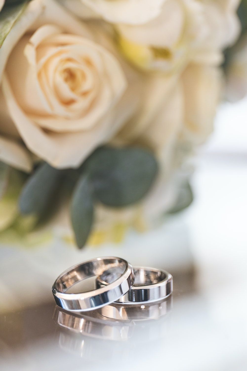 Two silver wedding rings on a white surface with a bouquet of white roses in the background. - Wedding