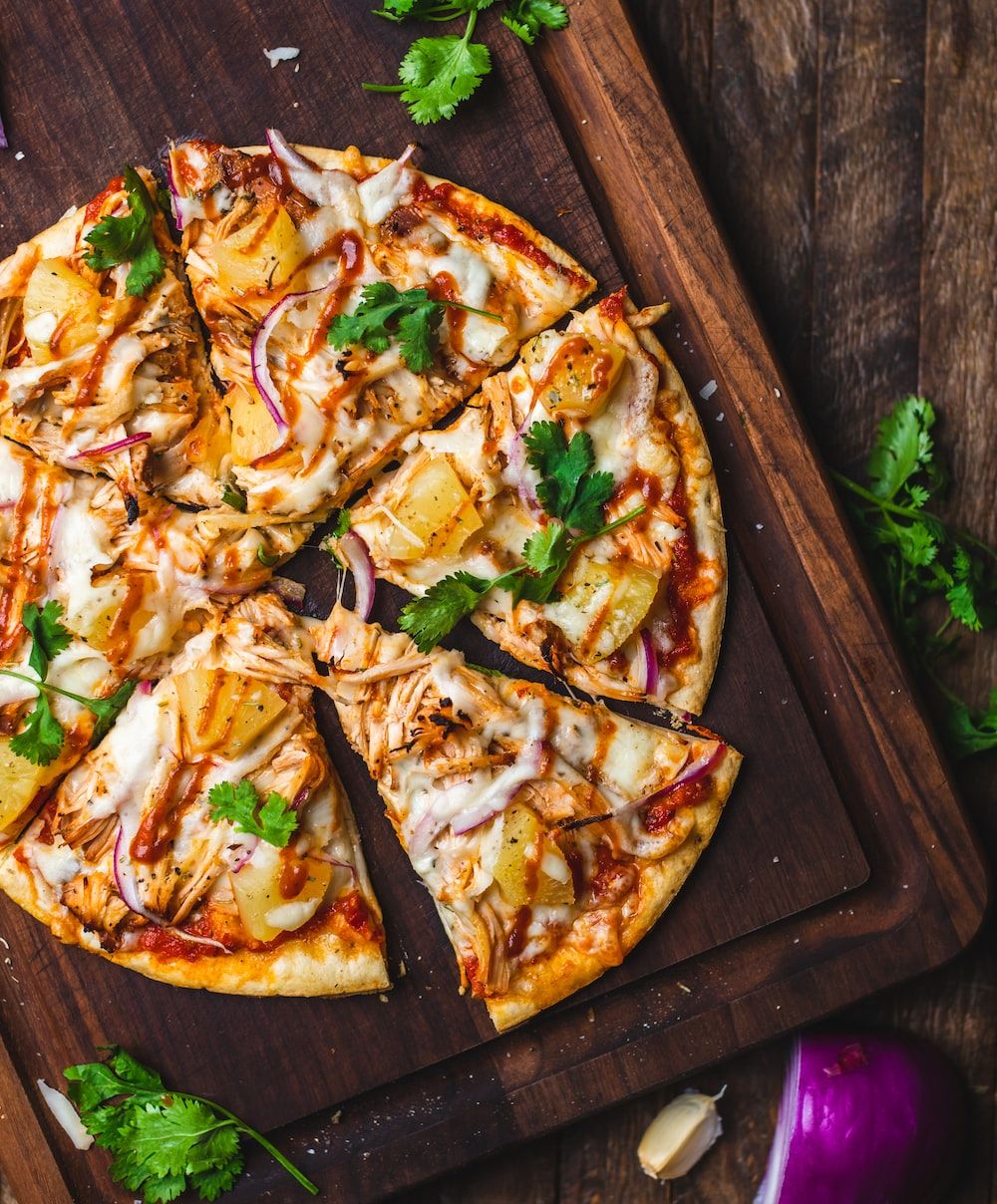 A pizza with chicken, pineapple, and red onions on a wooden cutting board. - Pizza