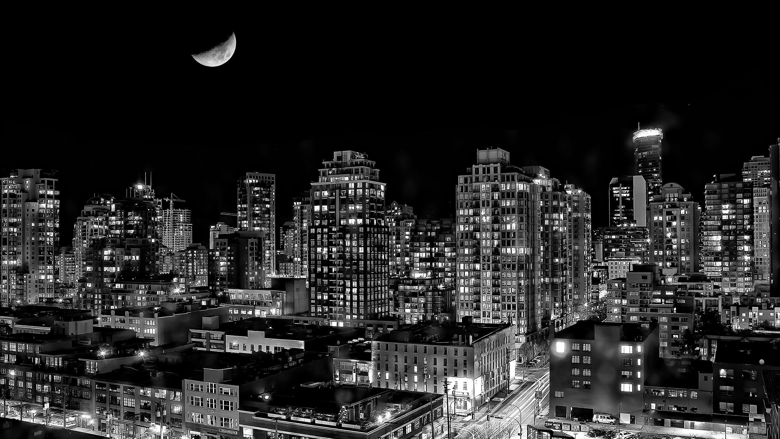 A black and white photo of a city at night with a half moon in the sky. - 2560x1440