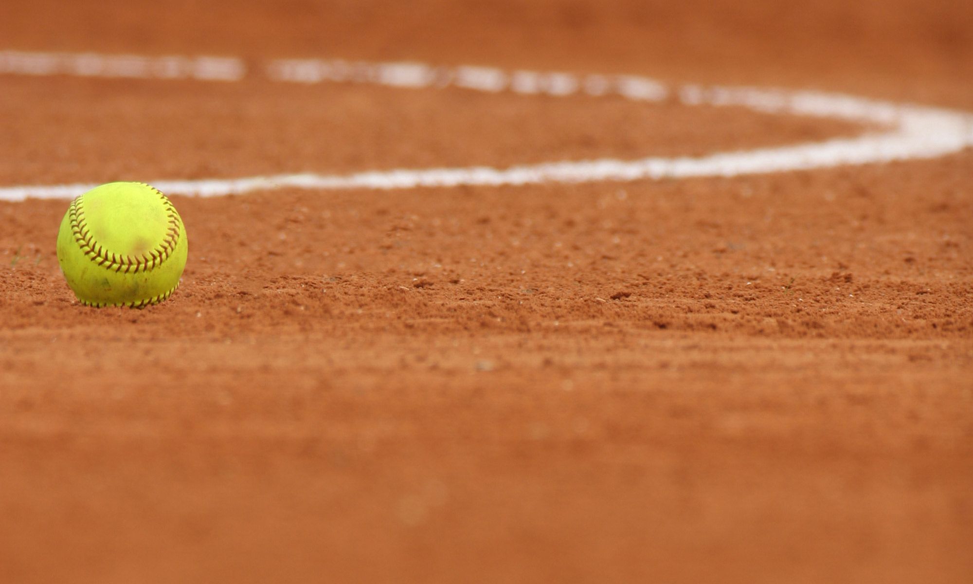 A yellow softball sits on the red clay of a softball field. - Softball