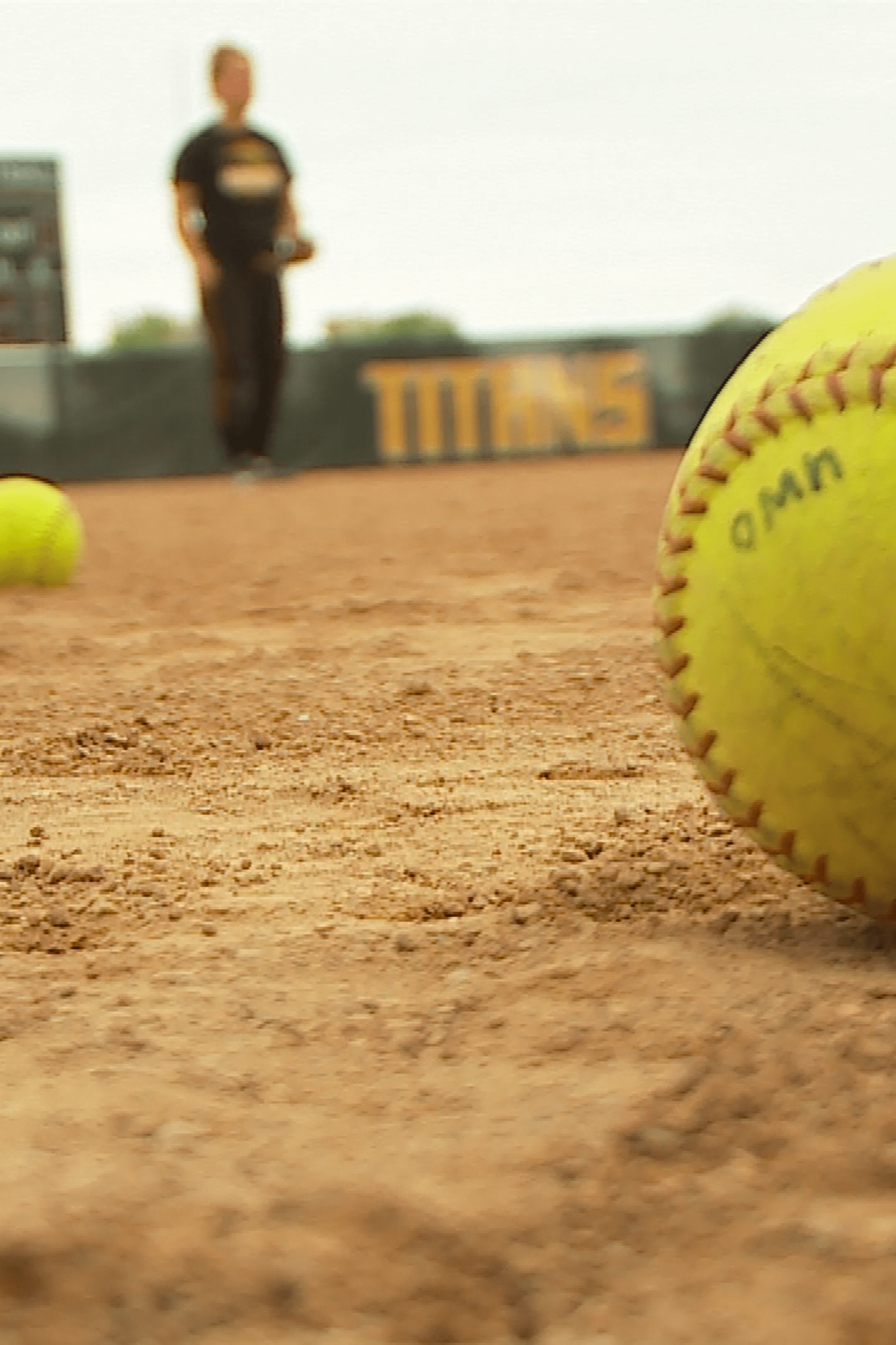 A woman standing on a field with a baseball and a ball behind her. - Softball