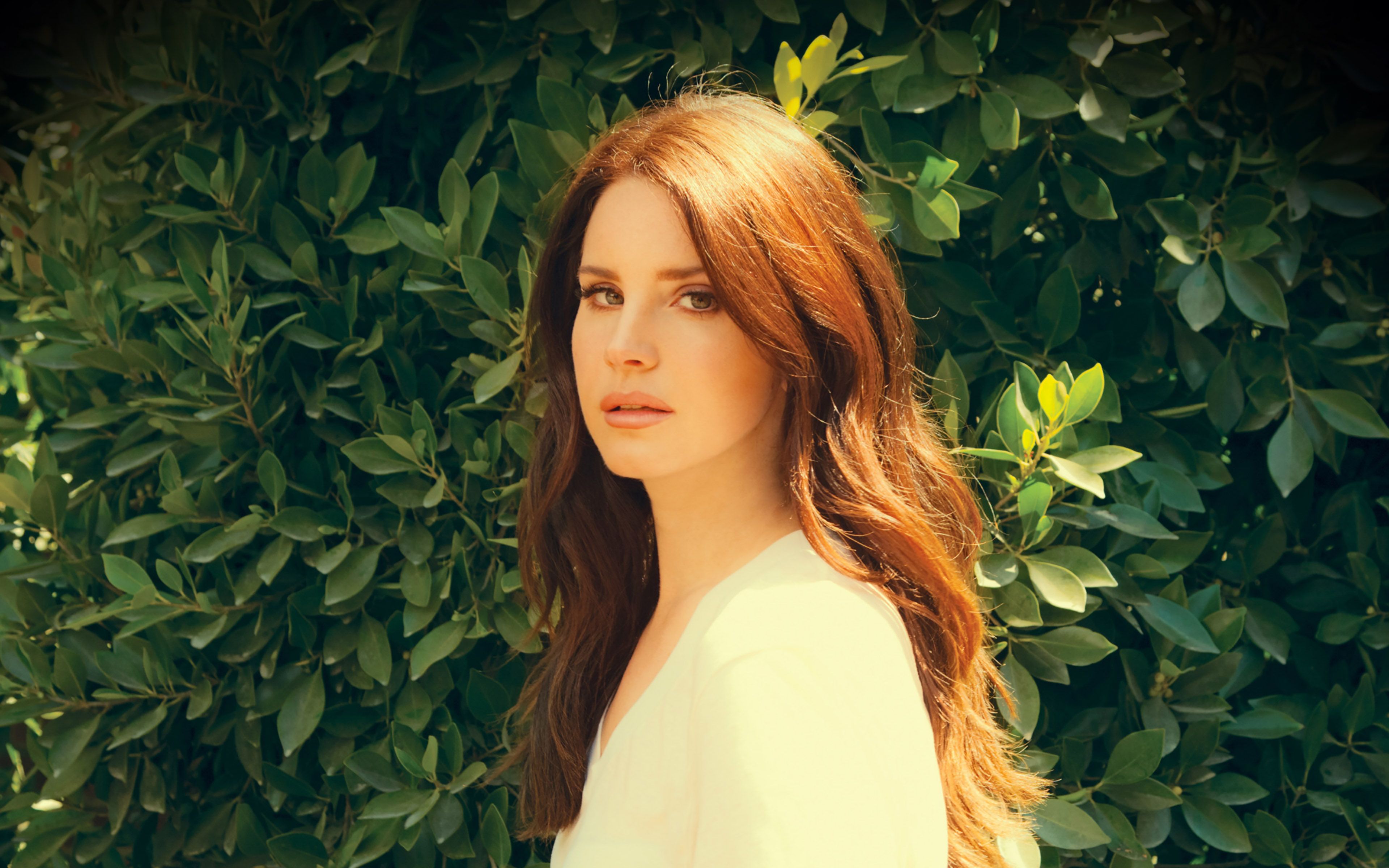 A woman with long red hair stands in front of a green bush. - Lana Del Rey