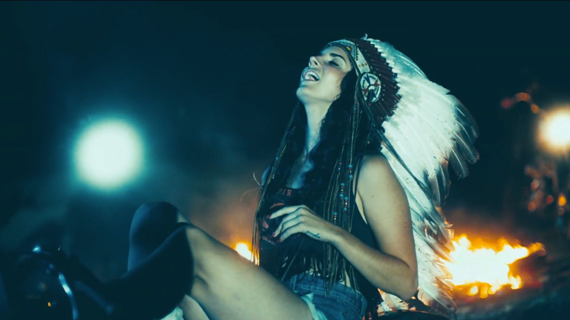 A woman wearing a Native American headdress sits on the ground laughing. - Lana Del Rey