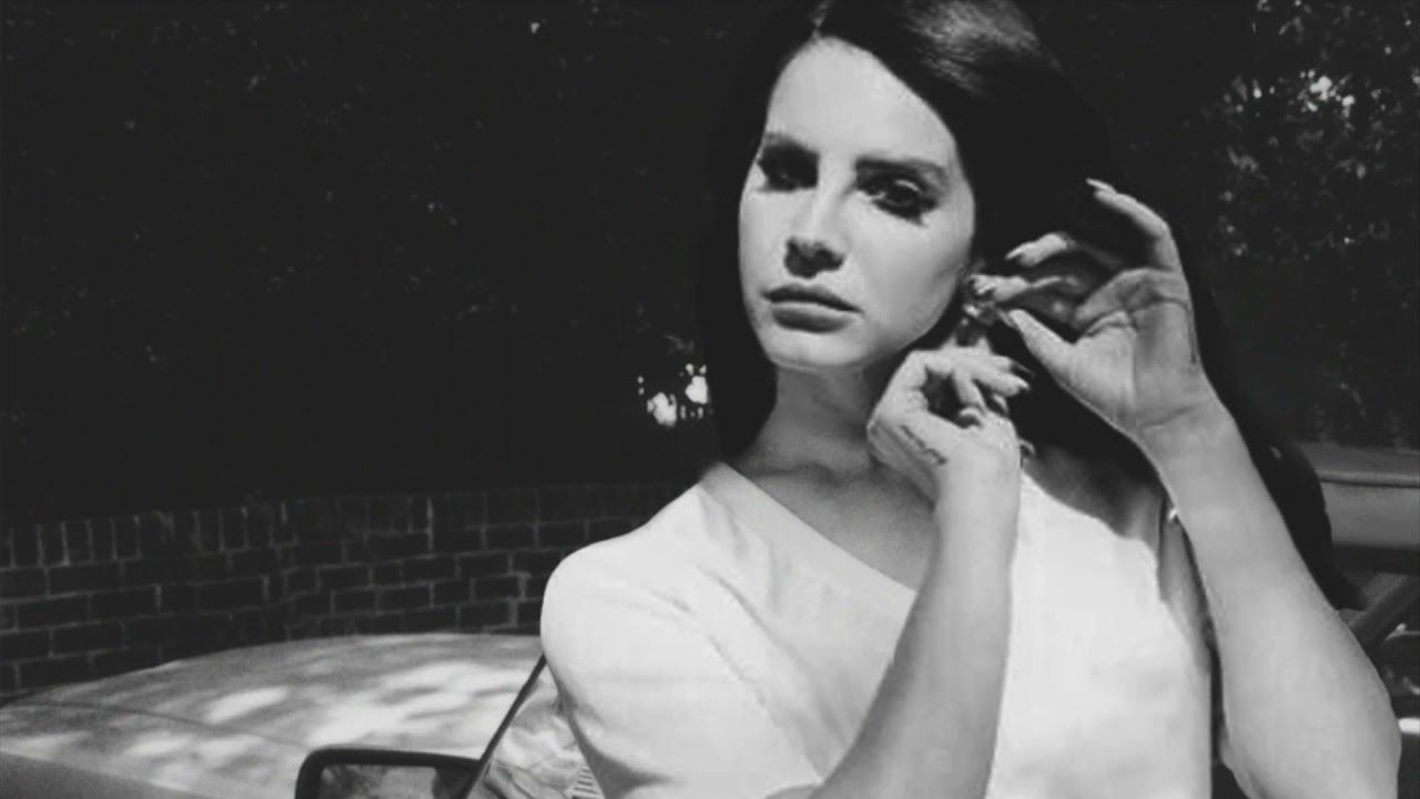 A woman in a white dress sits on a bench, looking into the camera. - Lana Del Rey