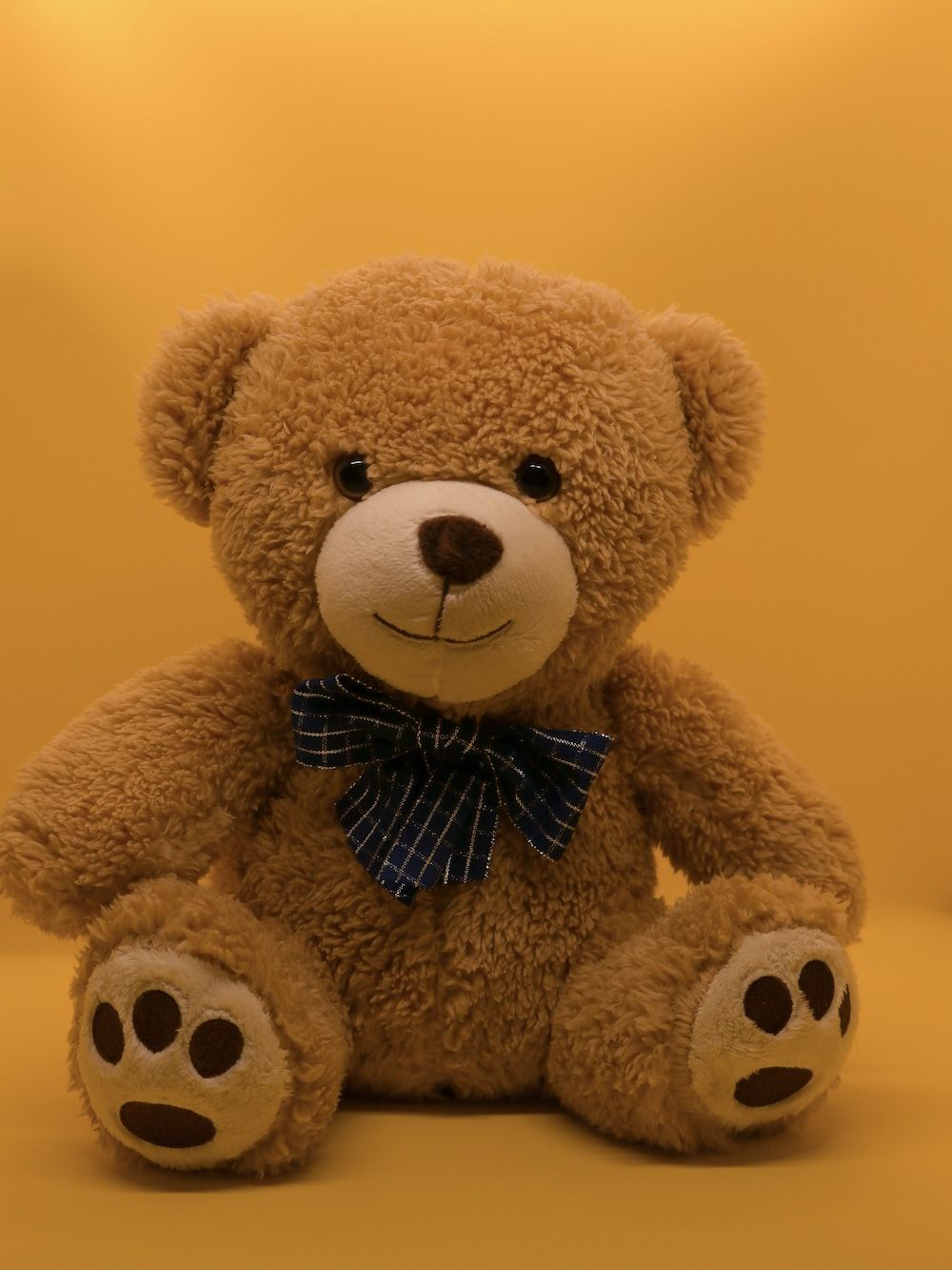 A brown teddy bear with a blue bow tie sitting on a yellow background - Teddy bear