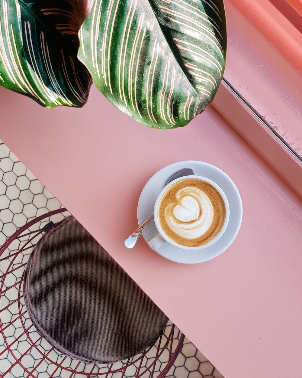 A cup of coffee sits on a pink table next to a green plant. - Coffee