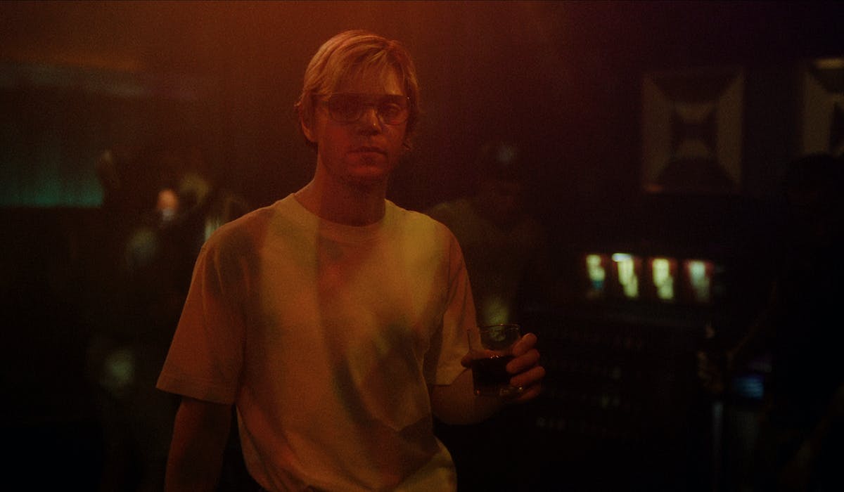 A young man with glasses and a drink stands in a darkened room. - Evan Peters