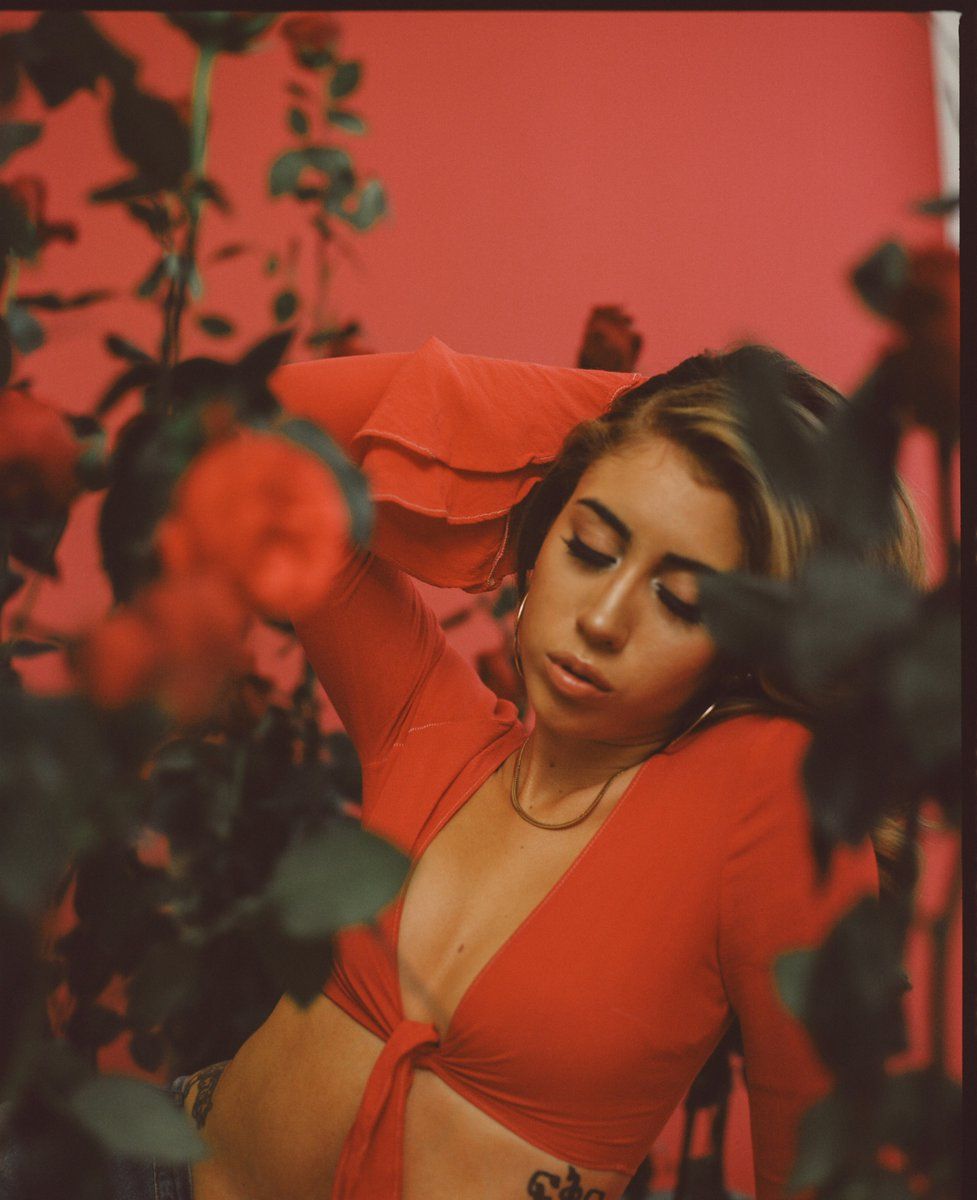 A woman in a red top stands in front of a red wall and red flowers. - Kali Uchis