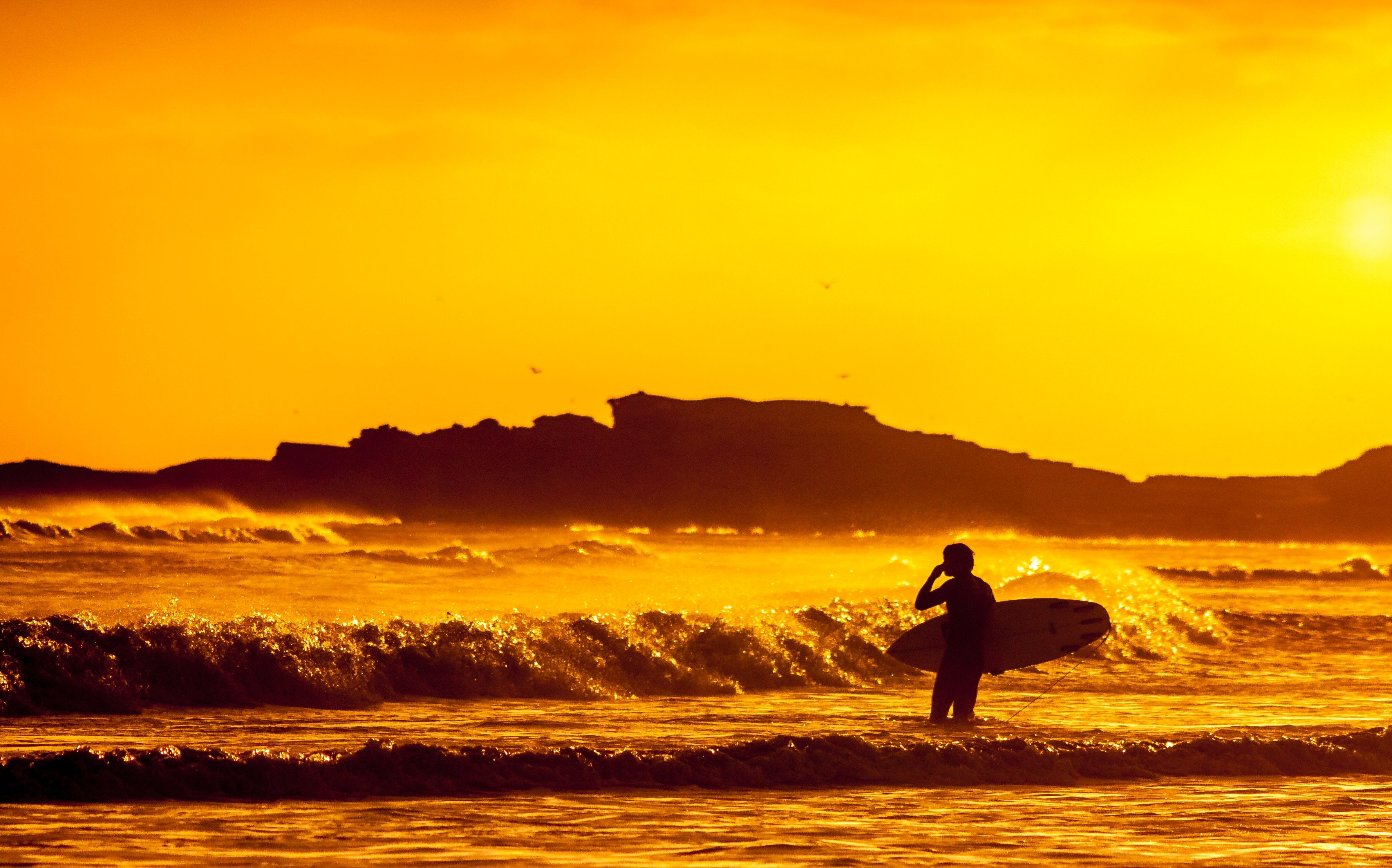 A surfer carries his board out of the ocean at sunset. - Surf