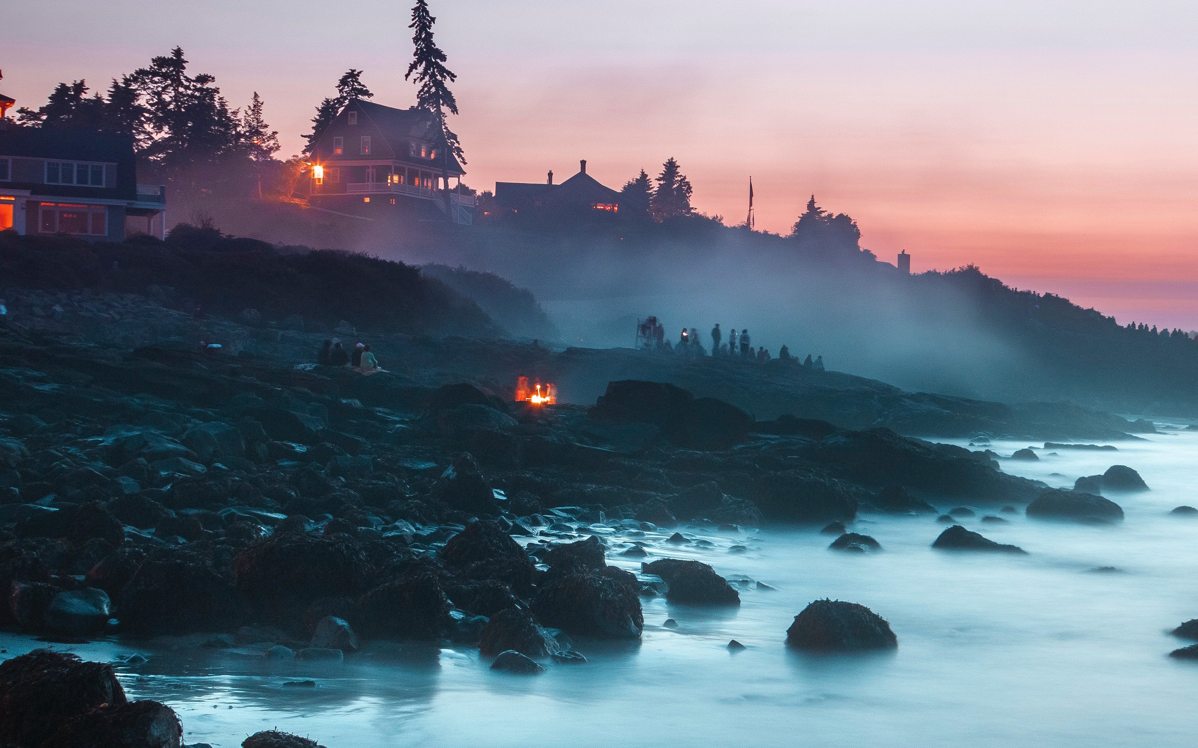 A group of people stand on a rocky beach at dusk, surrounded by mist. - MacBook