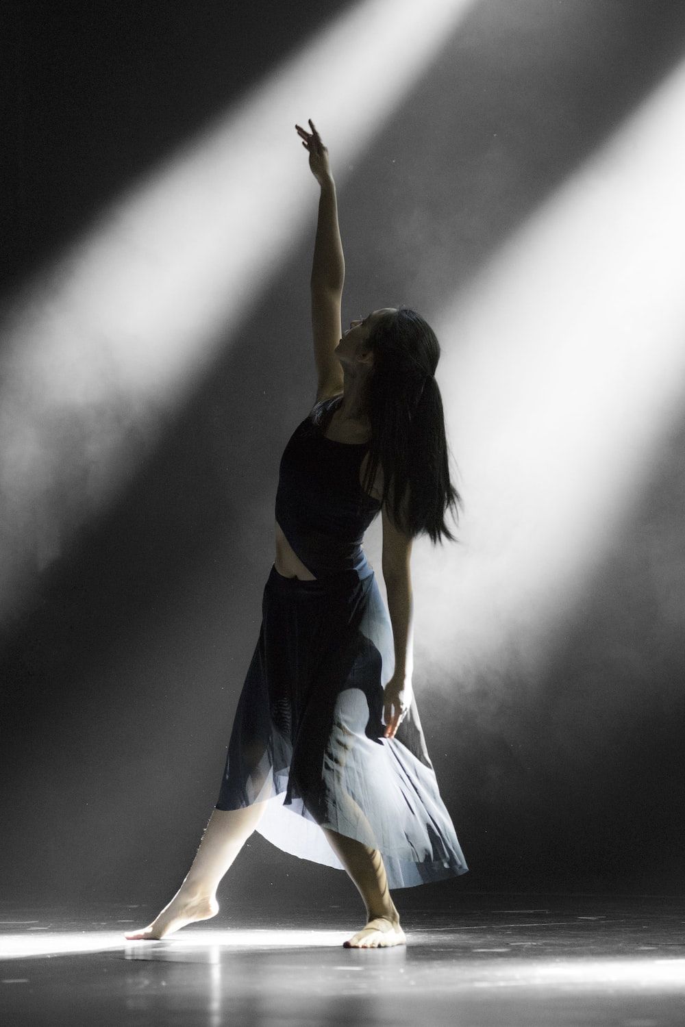A woman in a black top and flowing skirt dances on stage in a shaft of light. - Dance