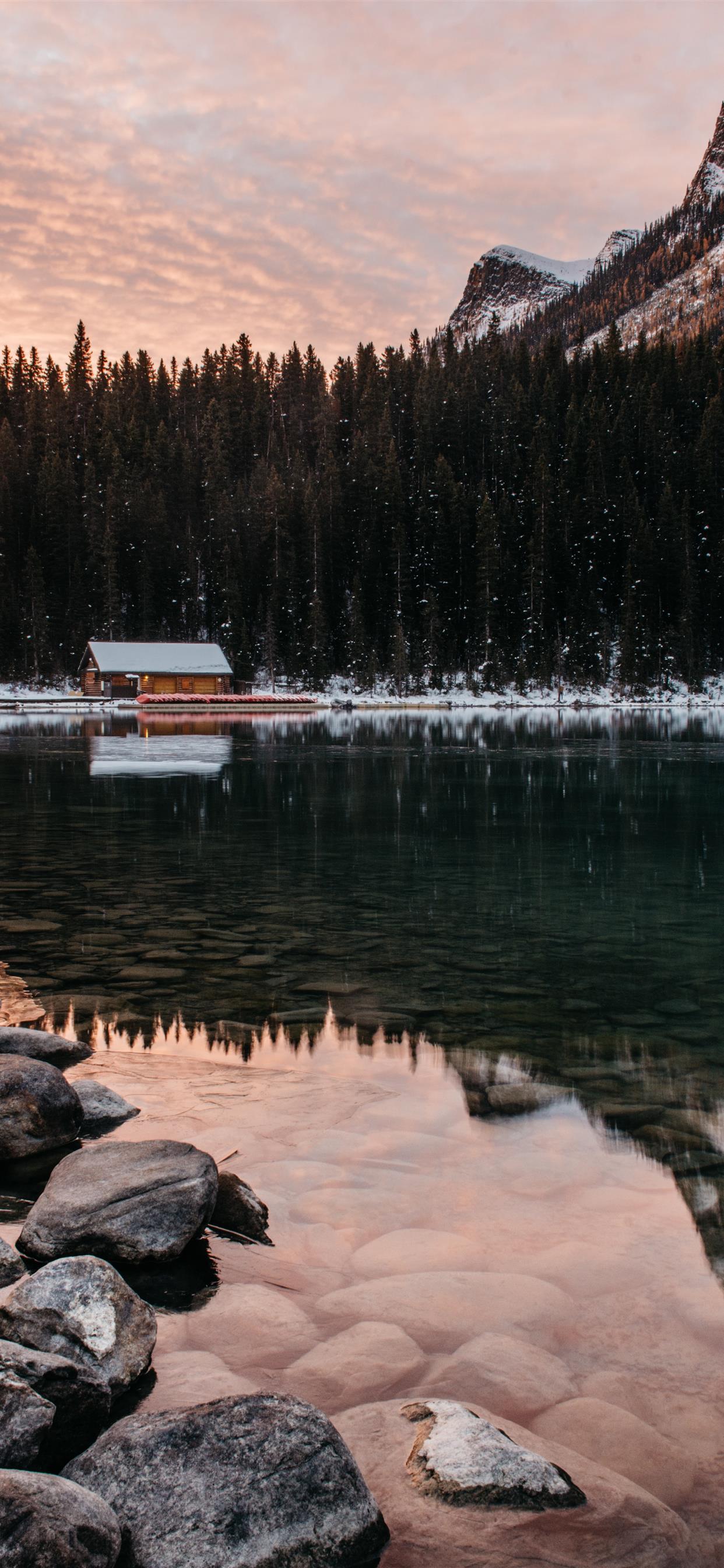 A small house sits on a lake surrounded by trees and mountains. - Winter, cozy, snow
