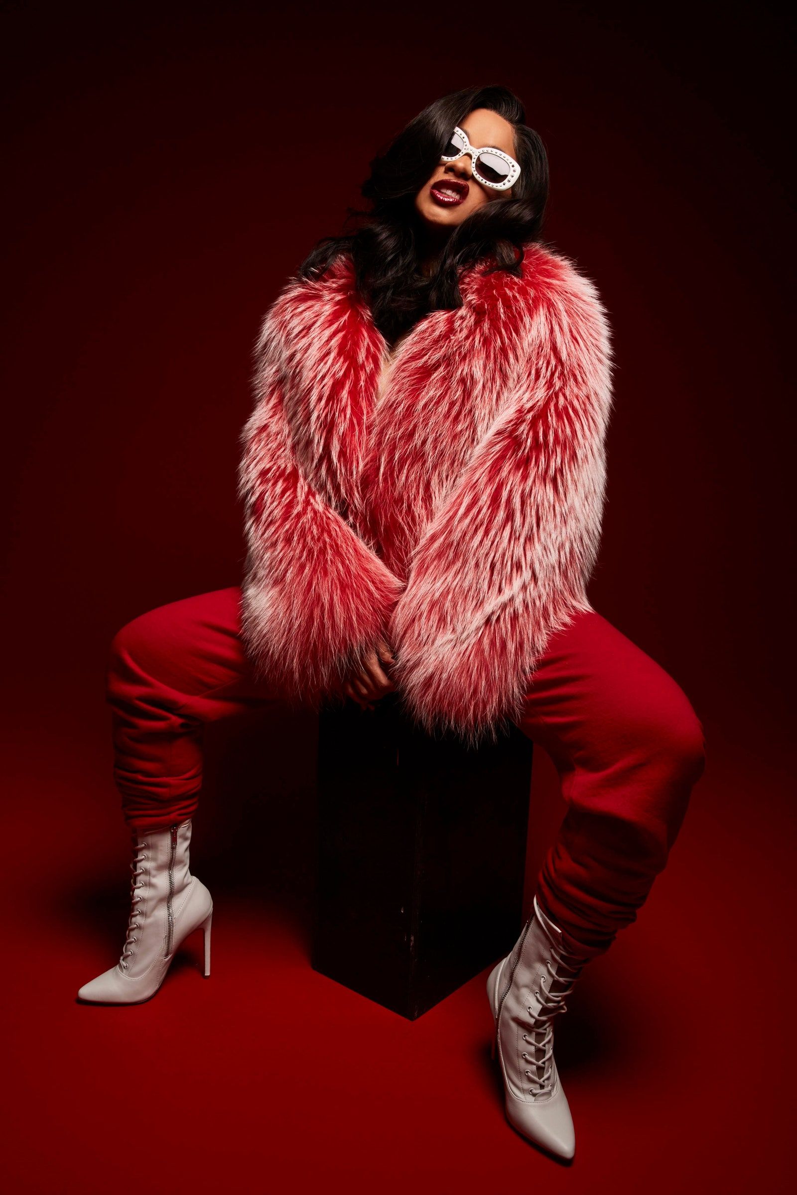 A woman in a red fur coat and white boots sits on a black box in front of a red background. - Cardi B