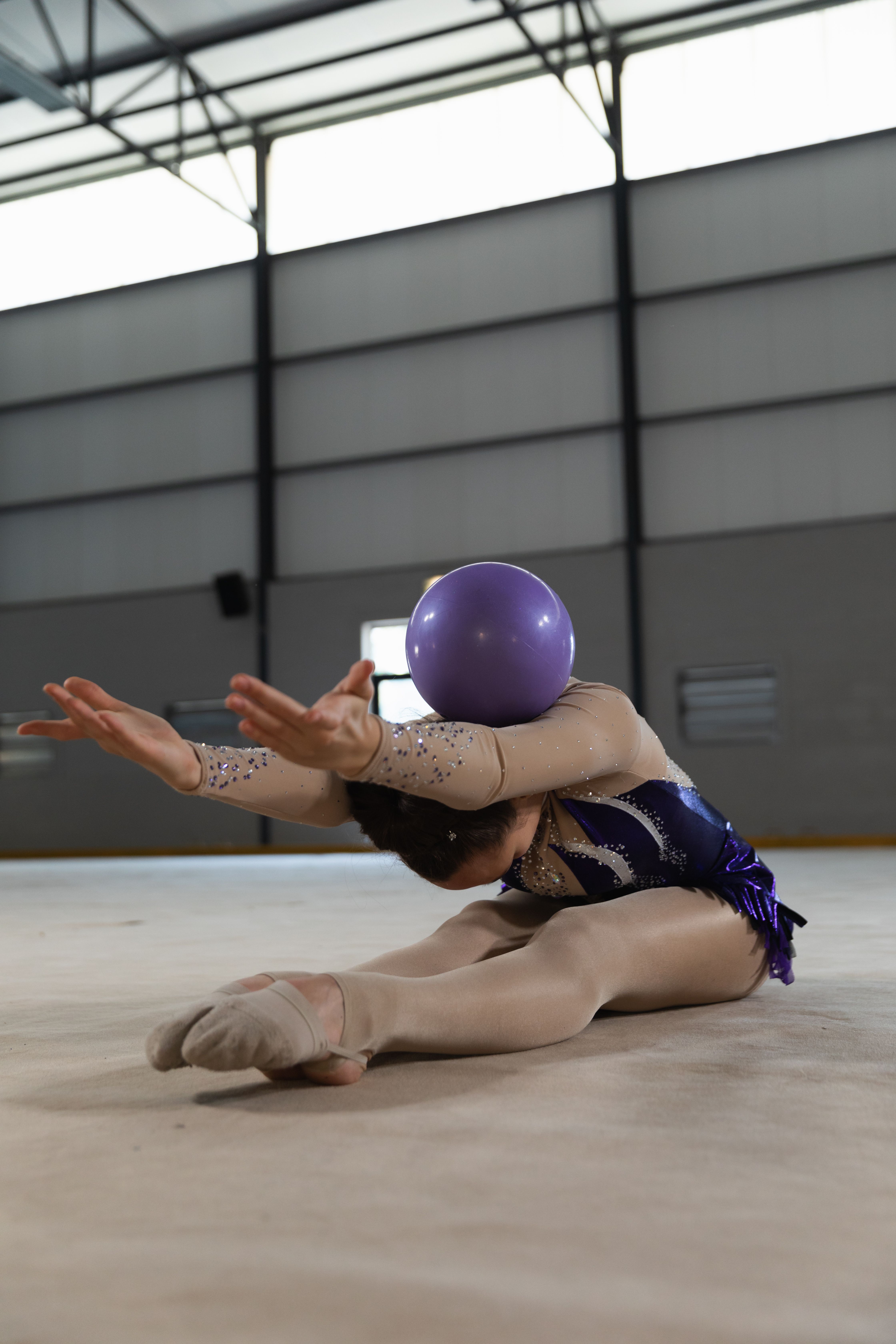 A gymnast stretches on the floor with a purple ball on her face - Gymnastics