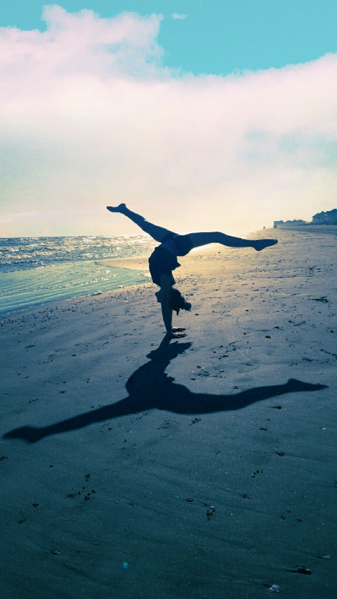 A person doing a handstand on the beach with the ocean in the background. The person is casting a long shadow on the sand. - Gymnastics