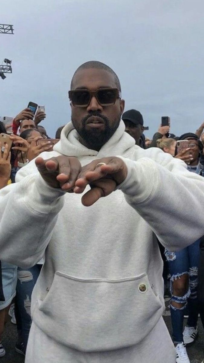 Kanye West in a white hoodie, with his hands out in front of him, and a crowd of people behind him. - Kanye West