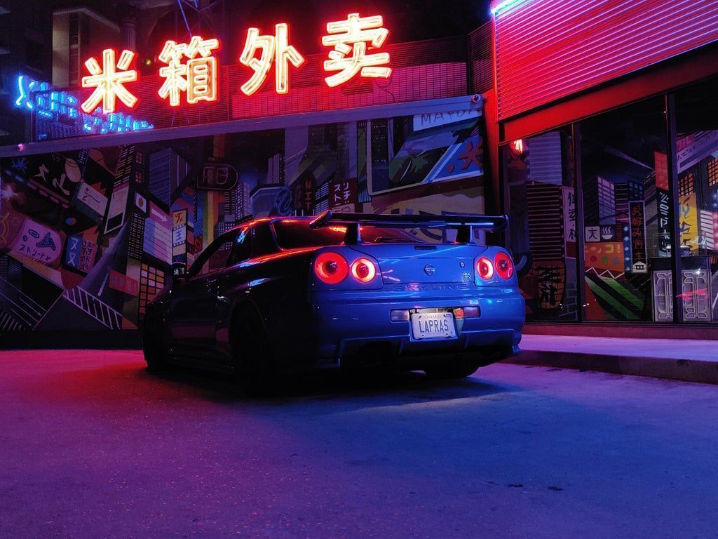 A blue car with red tail lights is parked in front of a store with red and blue neon lights. - Nissan Skyline, JDM, cars