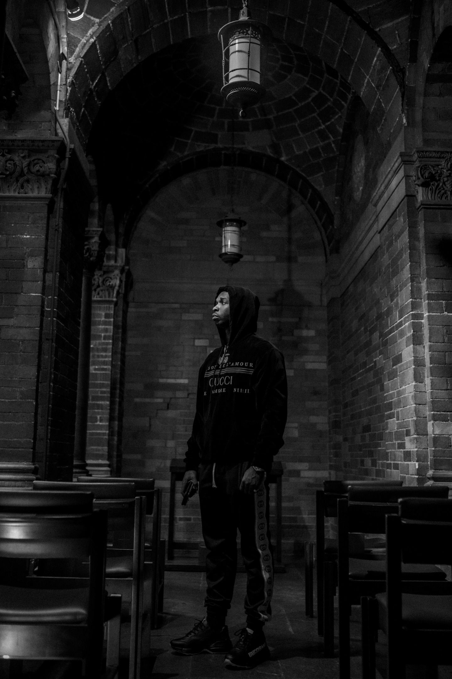 Grayscale photo of a man wearing a black hoodie standing in the middle of a church - Pop Smoke