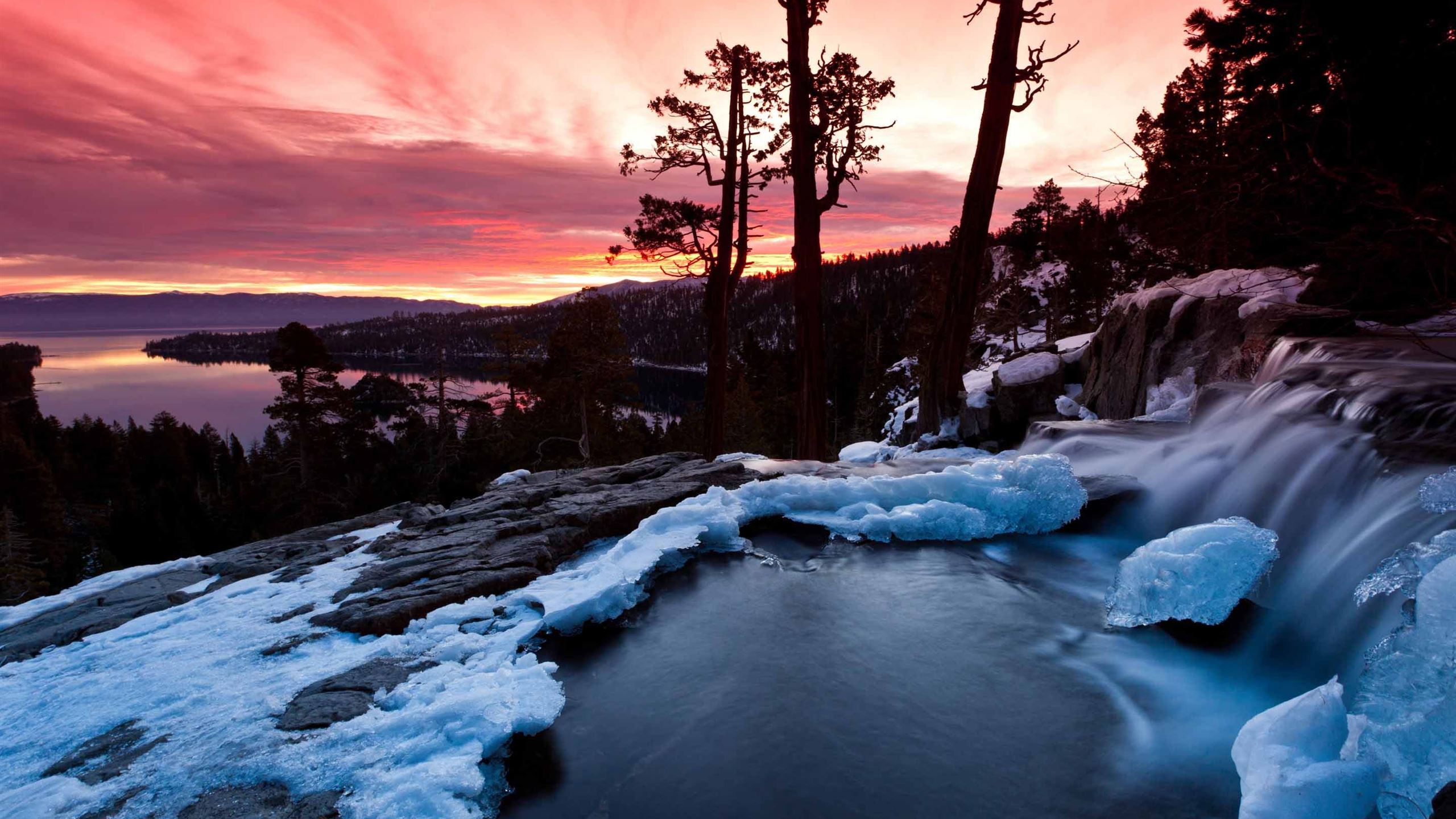 A beautiful winter sunset over Emerald Bay, Lake Tahoe, California with a frozen waterfall in the foreground and the silhouettes of trees in the background. - California