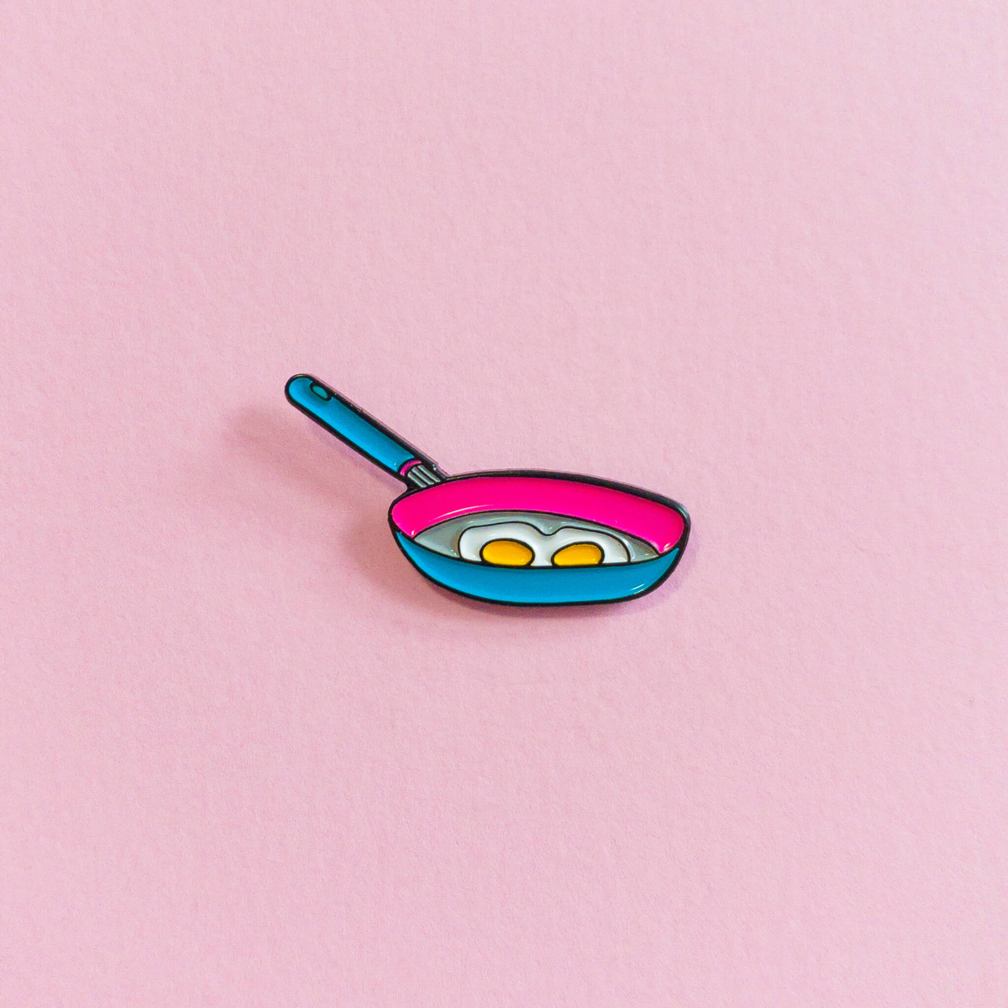 An enamel pin of two eggs frying in a pan, on a pink background. - Pansexual