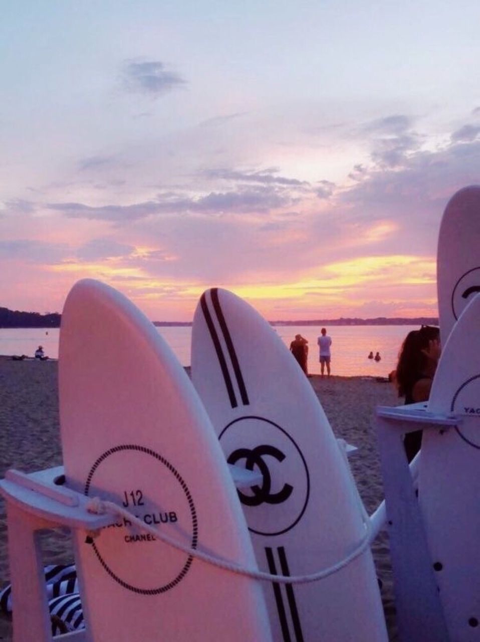 Surfboards are lined up on the beach with people in the water and a beautiful sunset in the background. - Surf