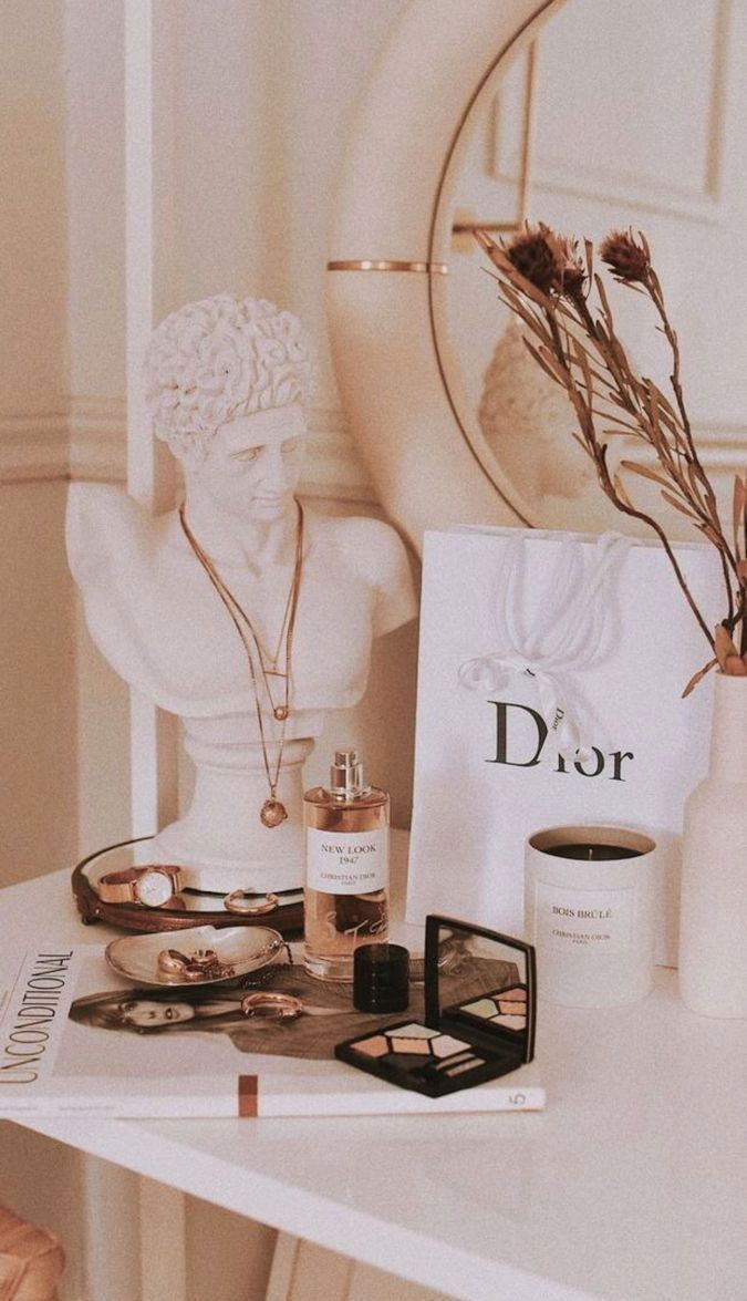 A table with a Dior bag, books, and a bust of a man. - Beige, cream, Dior, makeup, champagne