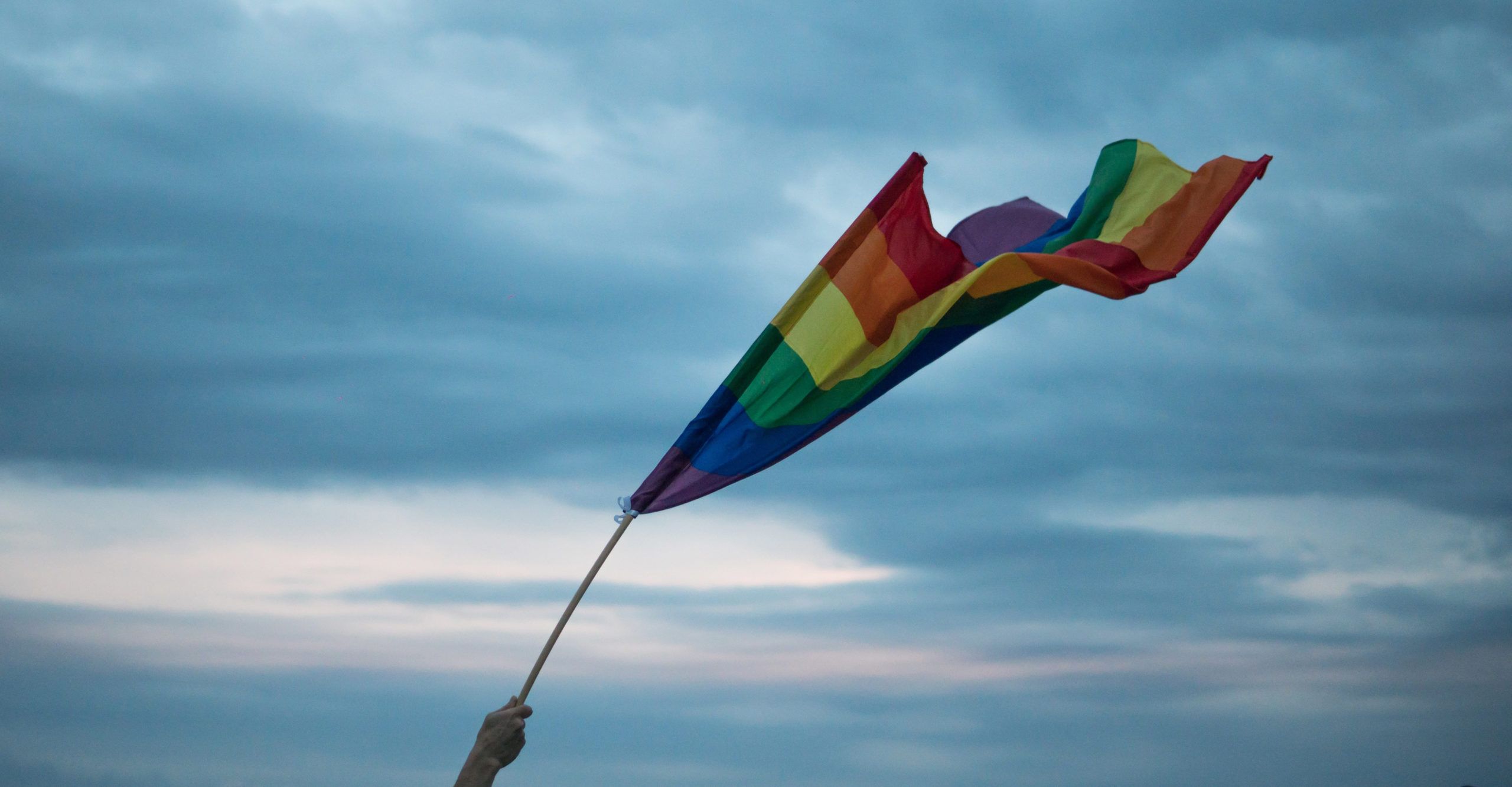 A hand holding a rainbow flag against a cloudy sky - Pride, gay
