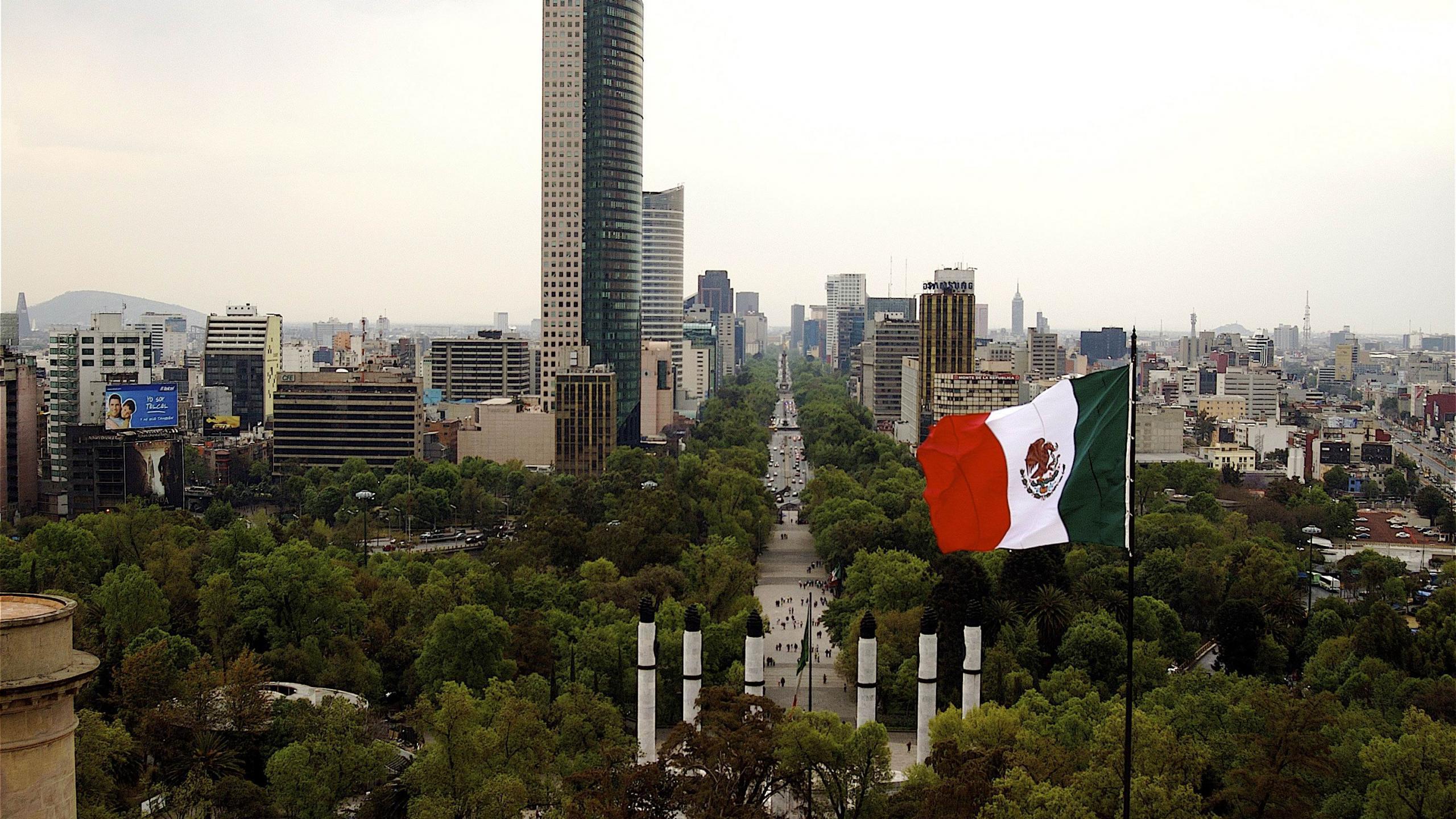 A Mexican flag waves in the wind in front of a cityscape. - Mexico