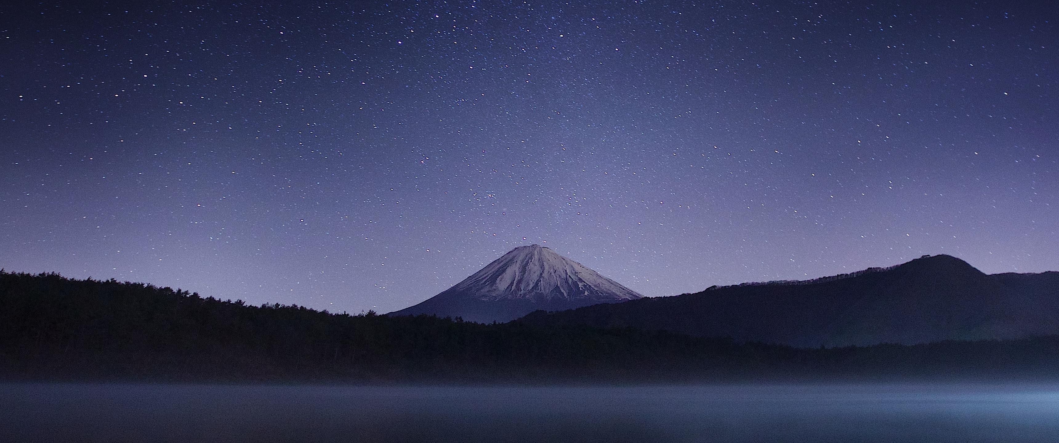 Mountain peak with snow on top, surrounded by a lake and a starry sky - 3440x1440